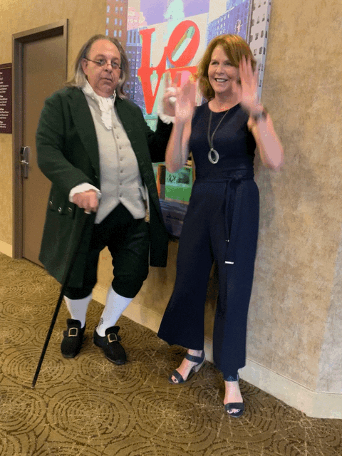 Two people standing indoors in front of a colorful background with the word 'LOVE' and a cityscape. The person on the left is dressed in period costume, wearing a green coat, white shirt, and black shoes with white socks. The person on the right is smiling, wearing a navy blue jumpsuit and high heels. They are both waving.
