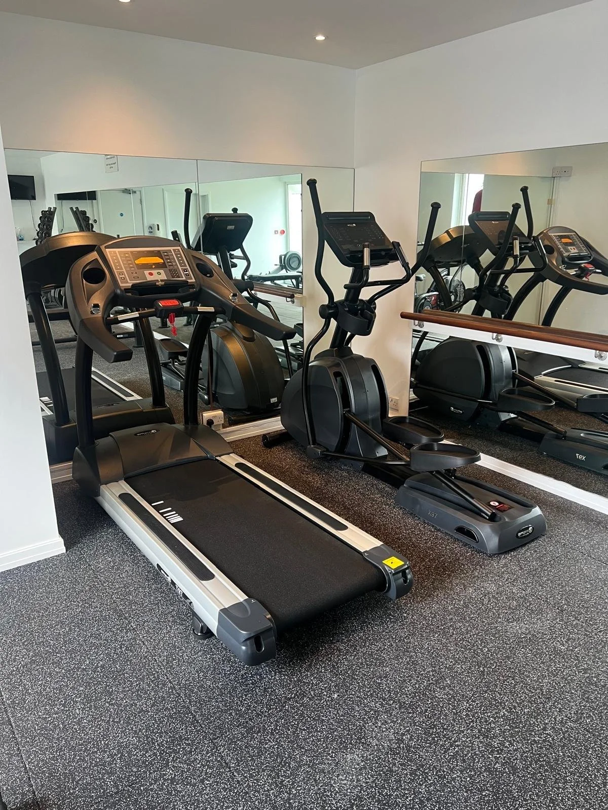 A gym room with various exercise equipment including a treadmill, an elliptical trainer, and other cardio machines, with a large mirror on the wall.