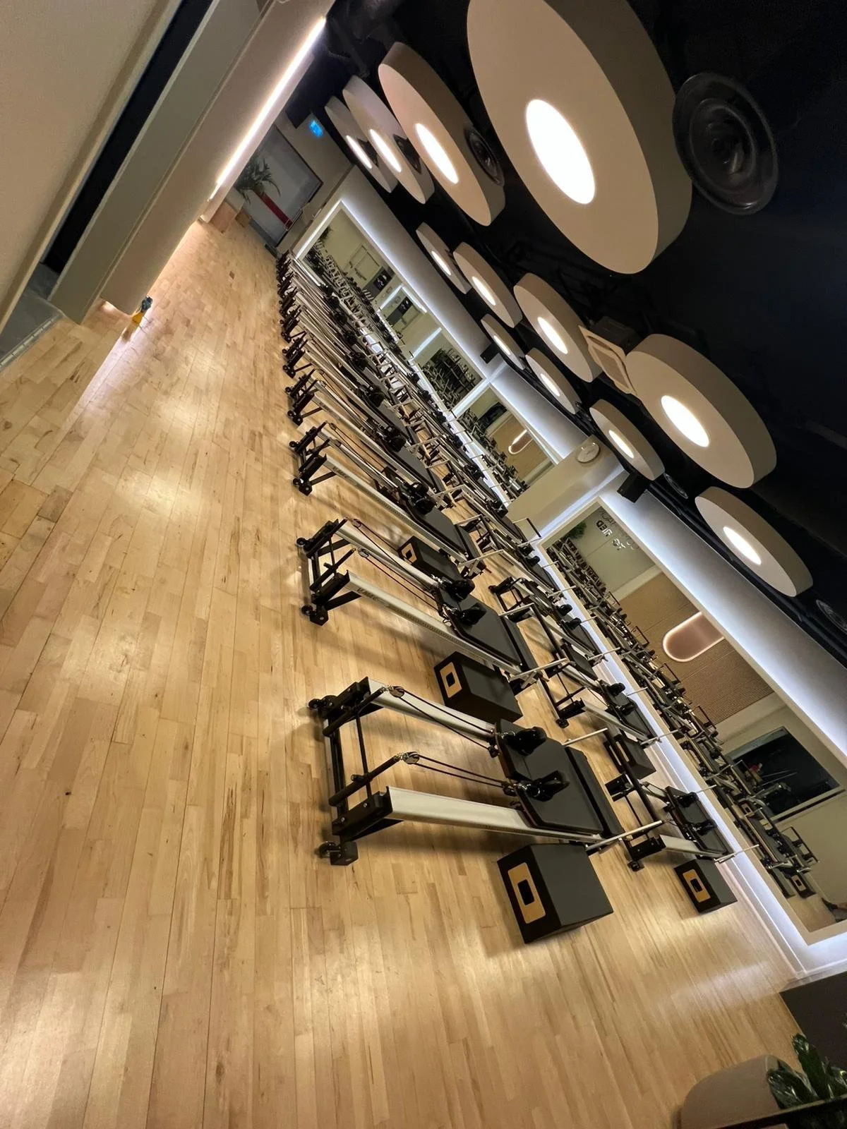 A spacious fitness studio with a row of Pilates reformer machines on a wooden floor, mirrored walls, and modern ceiling lights.