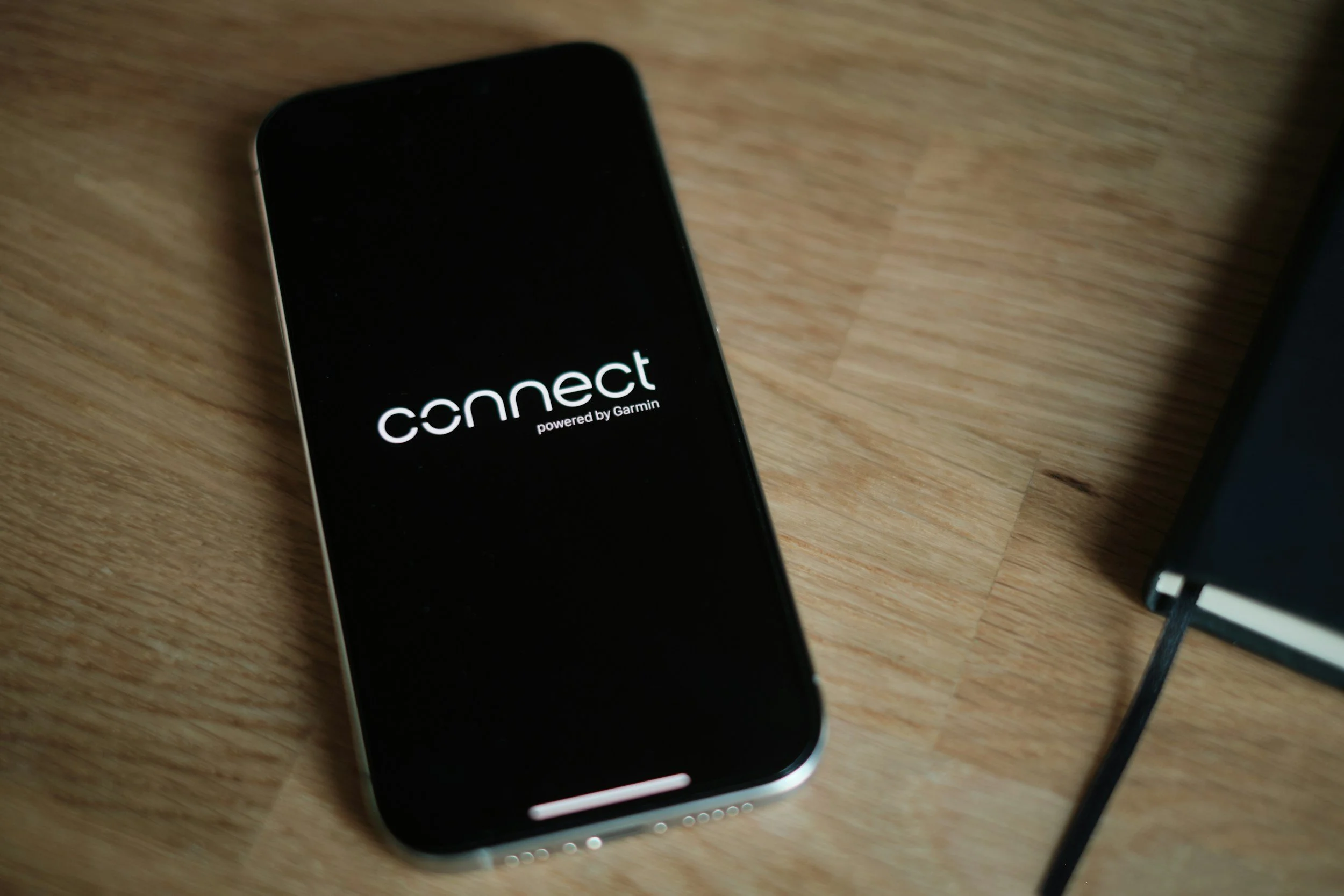A smartphone with the screen displaying the word 'connect' powered by Garmin, placed on a wooden surface next to a black notebook.