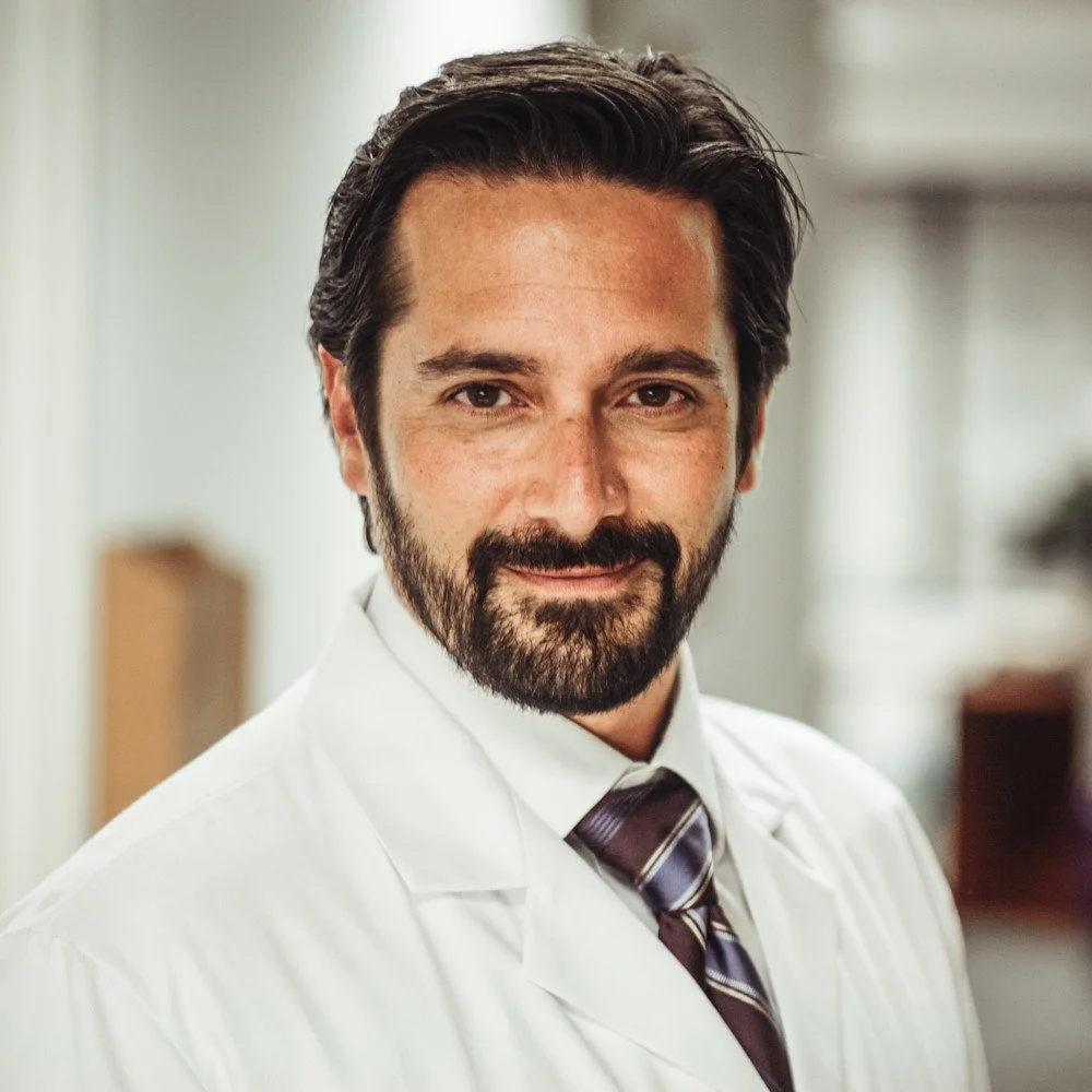 A man with dark hair and a beard wearing a white lab coat and a striped tie, smiling in a professional setting.