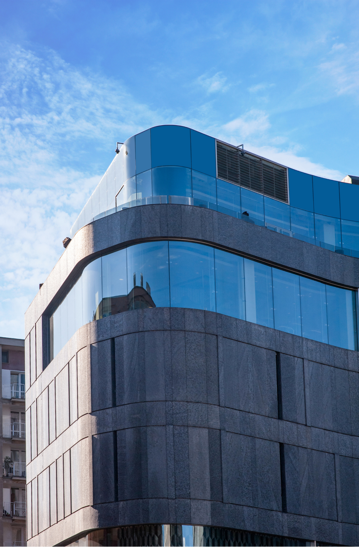 Modern multi-story building with curved glass windows reflecting the sky and clouds, and textured concrete exterior.