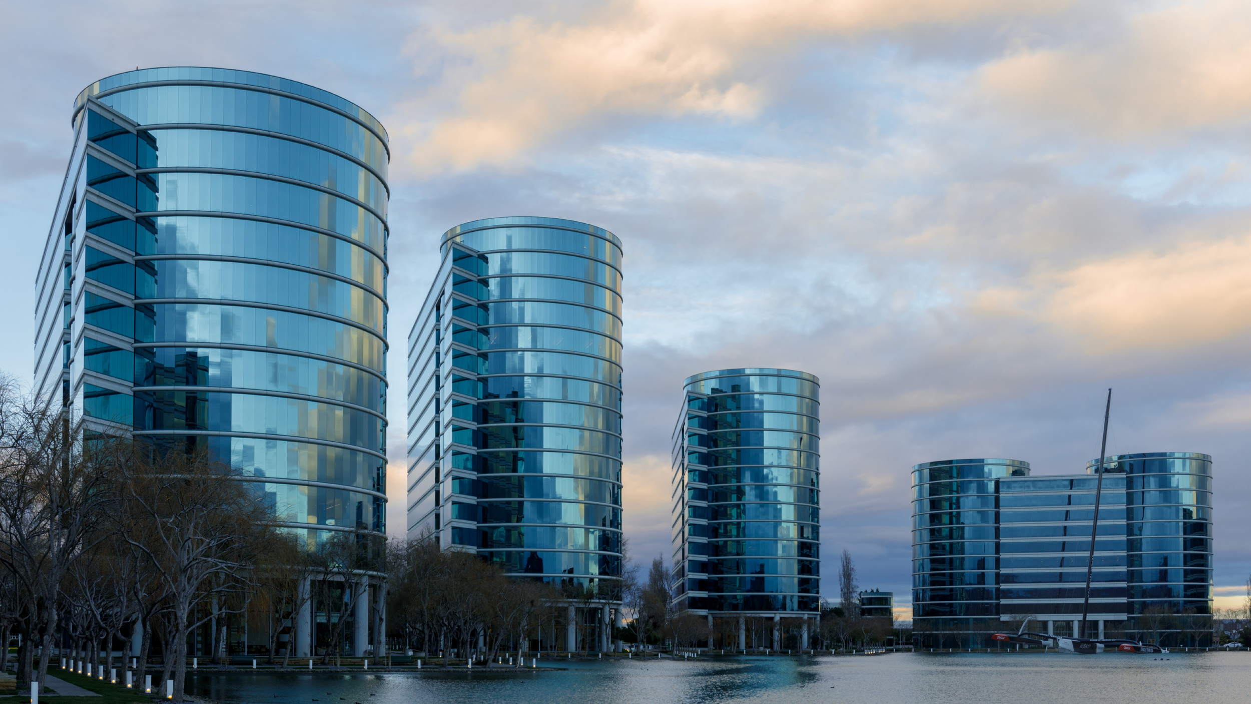 Modern glass high-rise buildings adjacent to a body of water with leafless trees along the shoreline, under a cloudy sky.