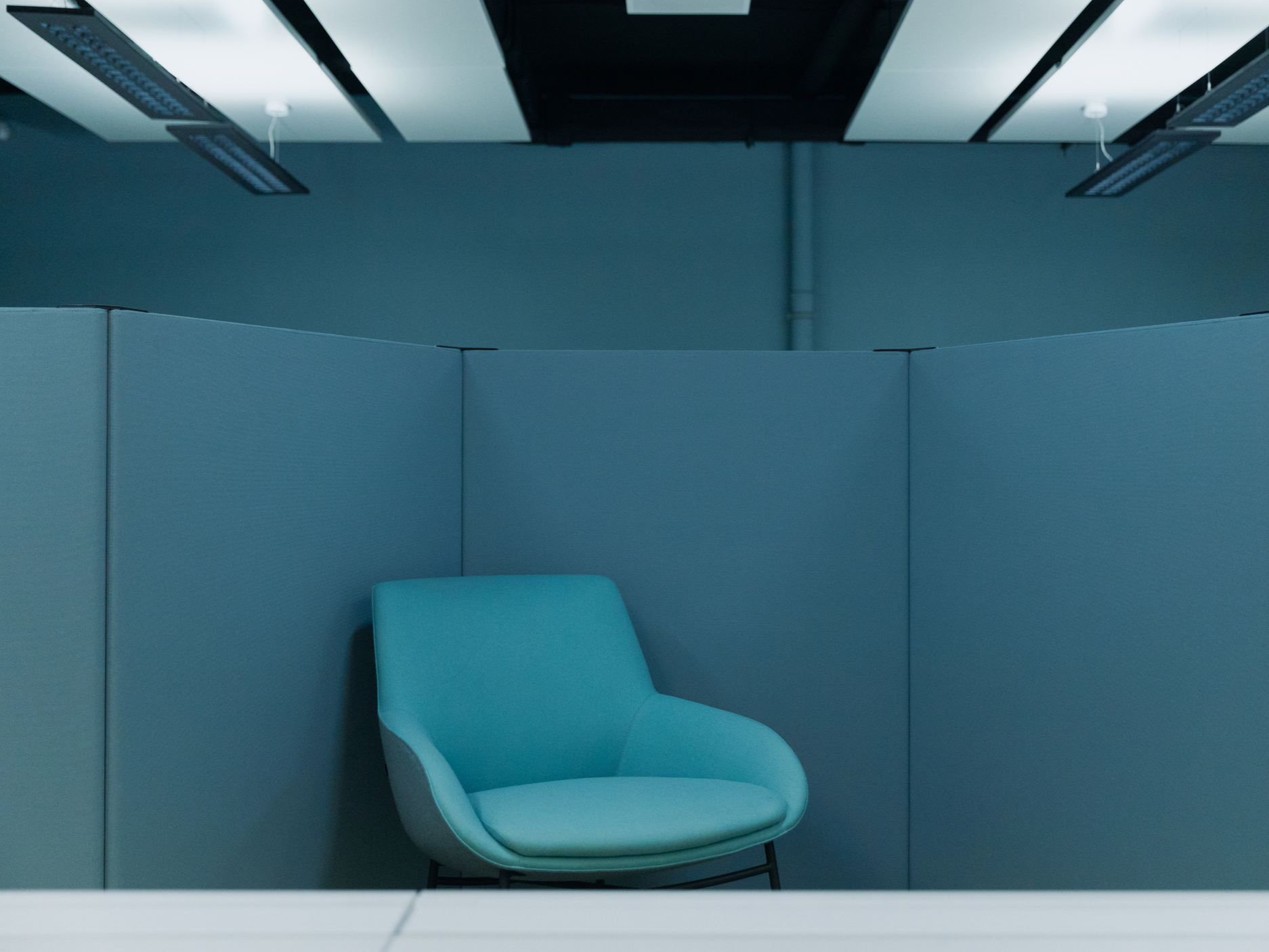 A blue office chair in a cubicle with blue partition walls, fluorescent ceiling lights, and a ceiling pipe.