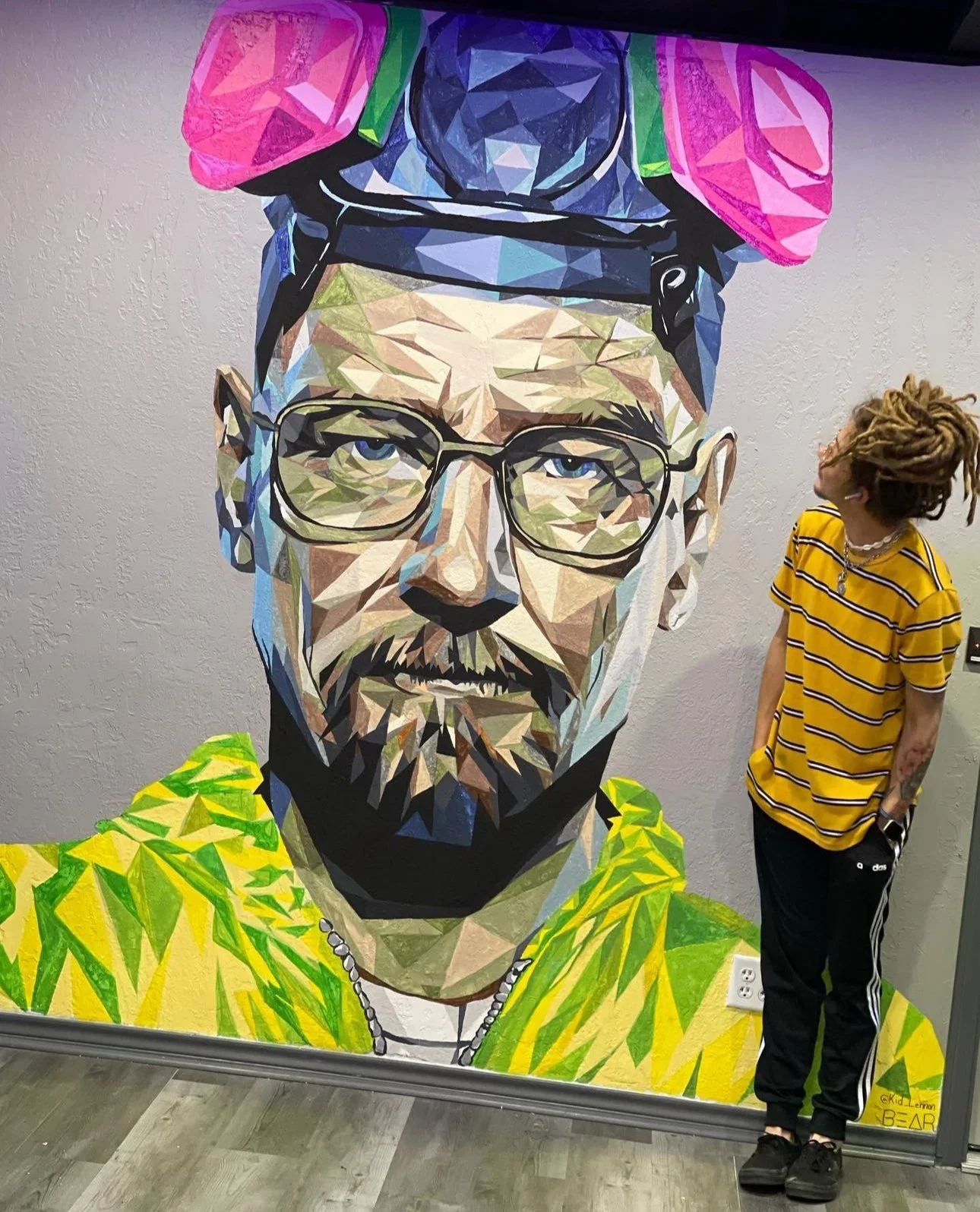A person with dreadlocks, wearing a yellow striped shirt and black pants, looking at a large geometric mural of a man's face on a wall.