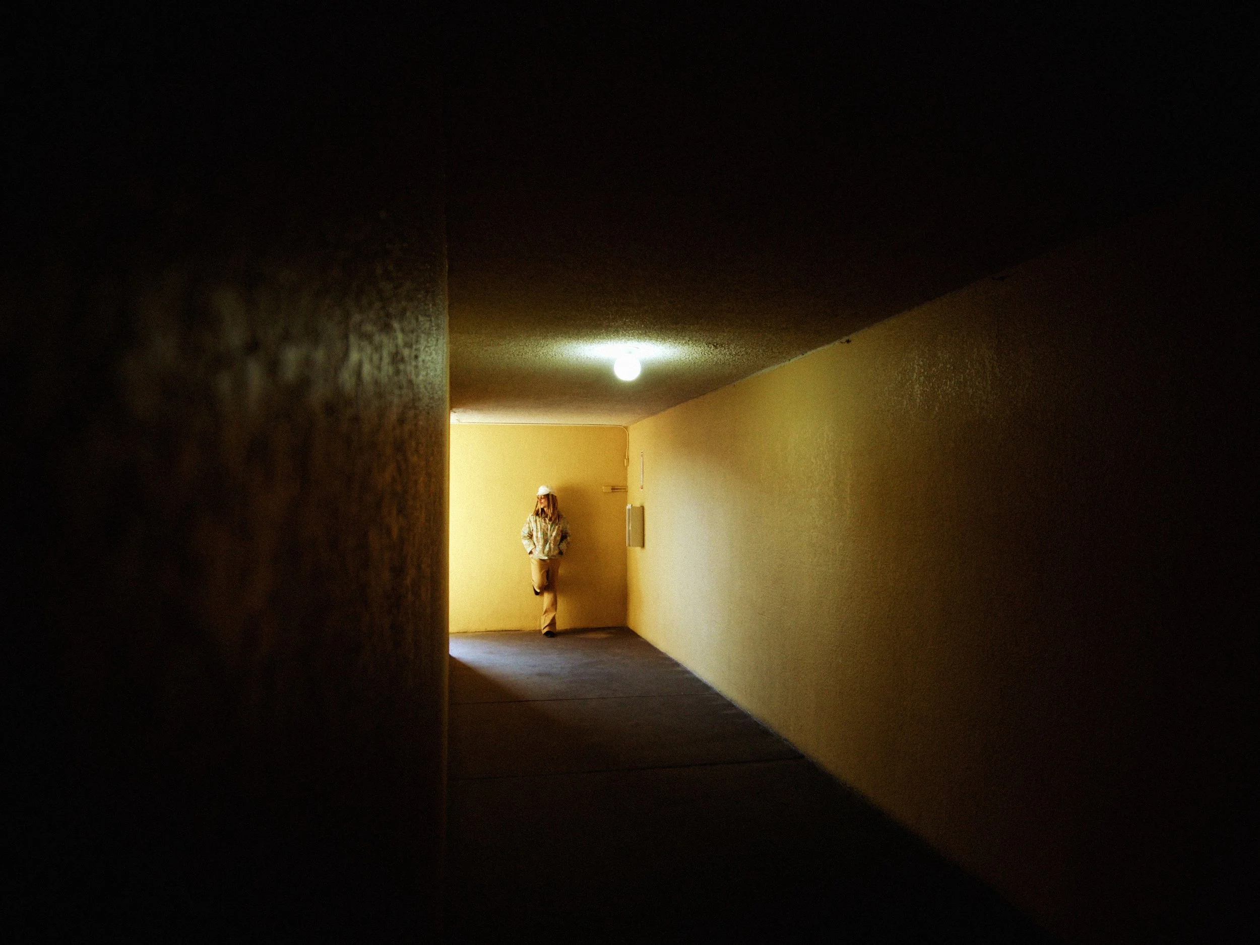 Person standing in a yellow corridor, leaning against the wall, wearing a hat and patterned shirt.