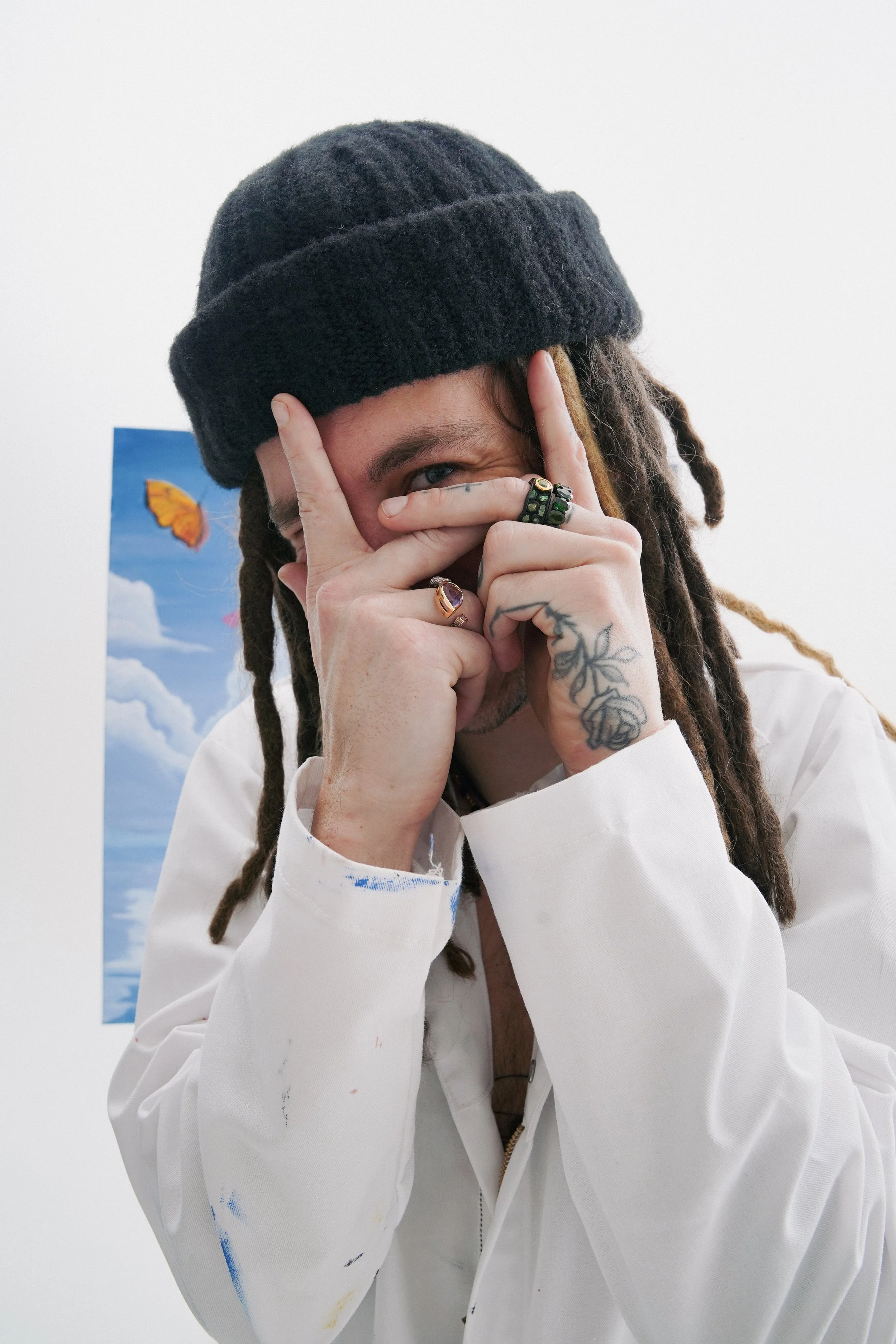 Person with dreadlocks wearing a black beanie and white smock, partially covering their face, with a tattoo on their hand, rings, and a colorful background with a cartoon butterfly.