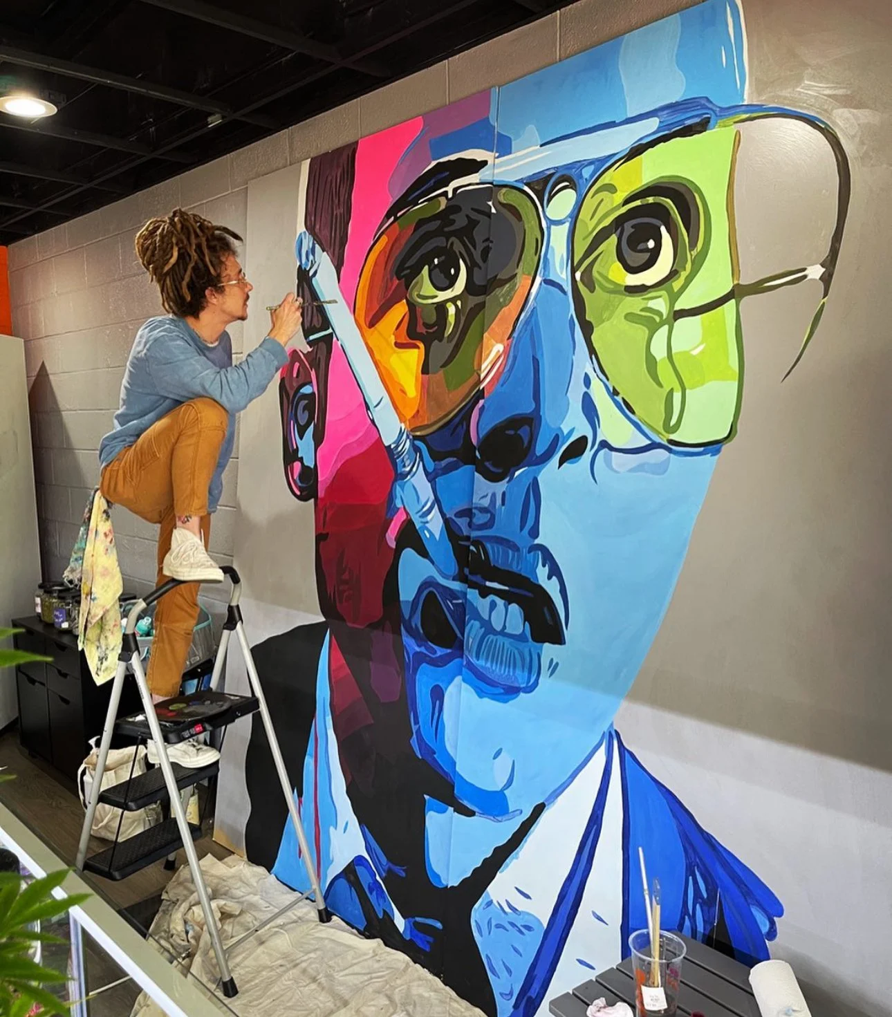 Artist working on a large colorful mural of a man's face with glasses, painted in a pop art style.