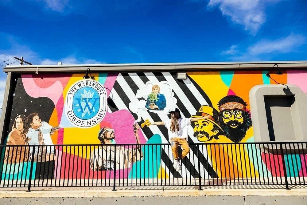 Colorful mural on a building featuring illustrations of people, two with beards and hats, and a woman in a white shirt. The mural also includes the logo of The Warehouse Dispensary and a person hanging from the railing in front of the mural.