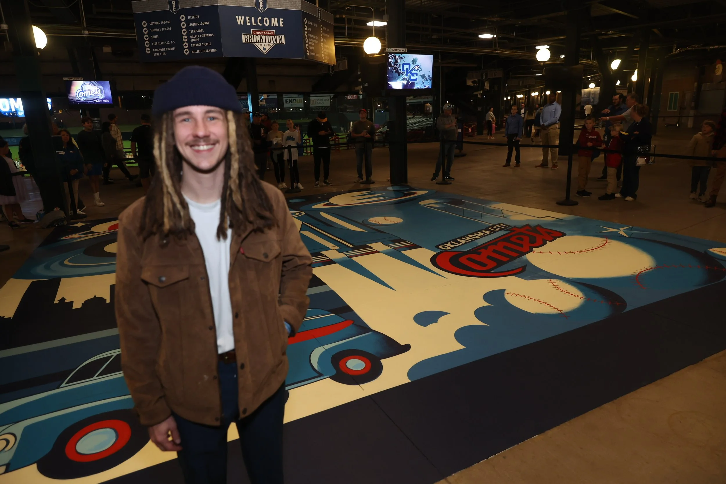 A young man with dreadlocks, wearing a brown jacket and a dark beanie, smiling in front of a painted basketball court on the floor at the Oklahoma City Thunder stadium. In the background, people are waiting in line and a large sign reads "Welcome Chickasaw Bricktown".