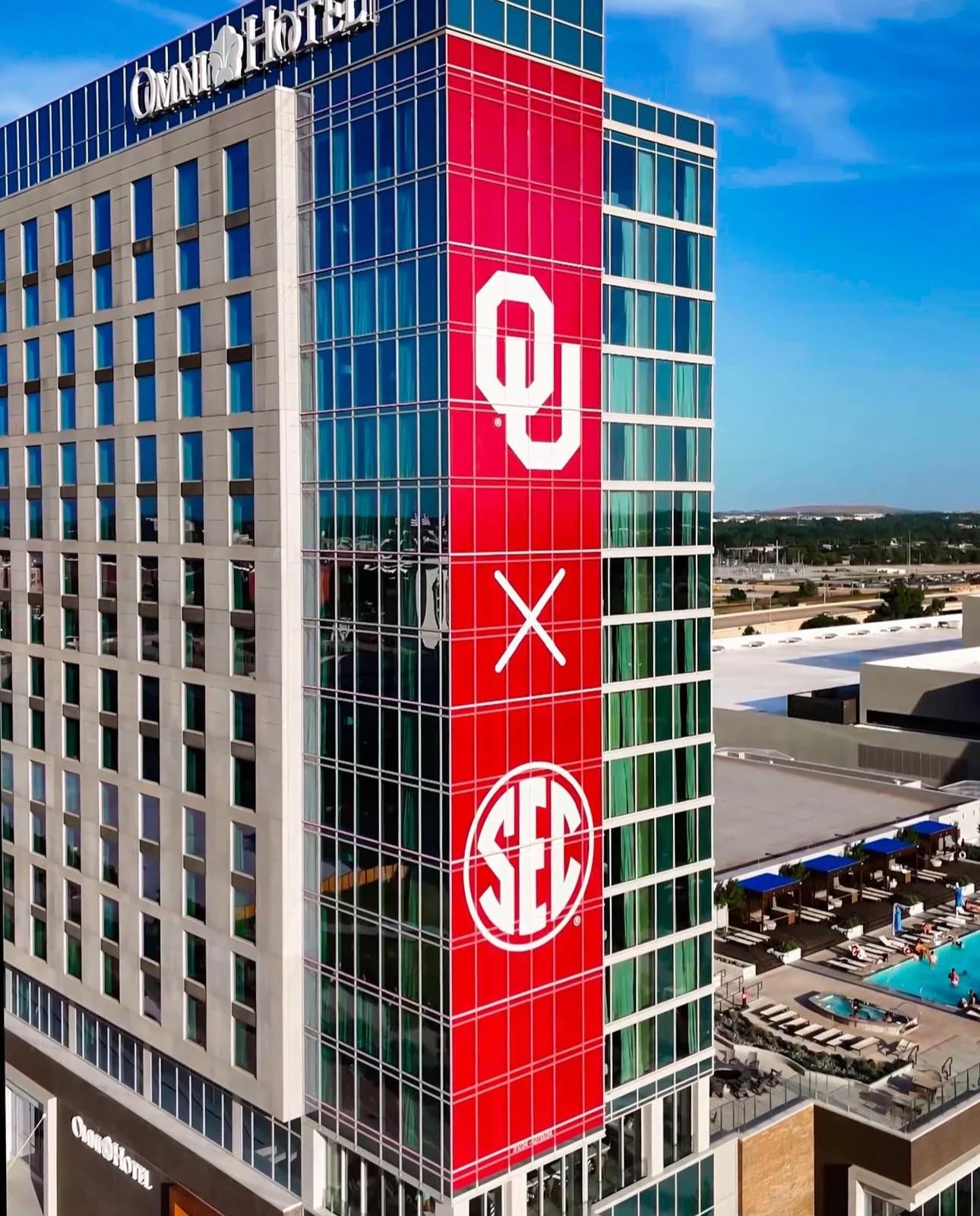 Tall building with a red vertical banner displaying the University of Oklahoma and SEC logos, with a pool area visible nearby.
