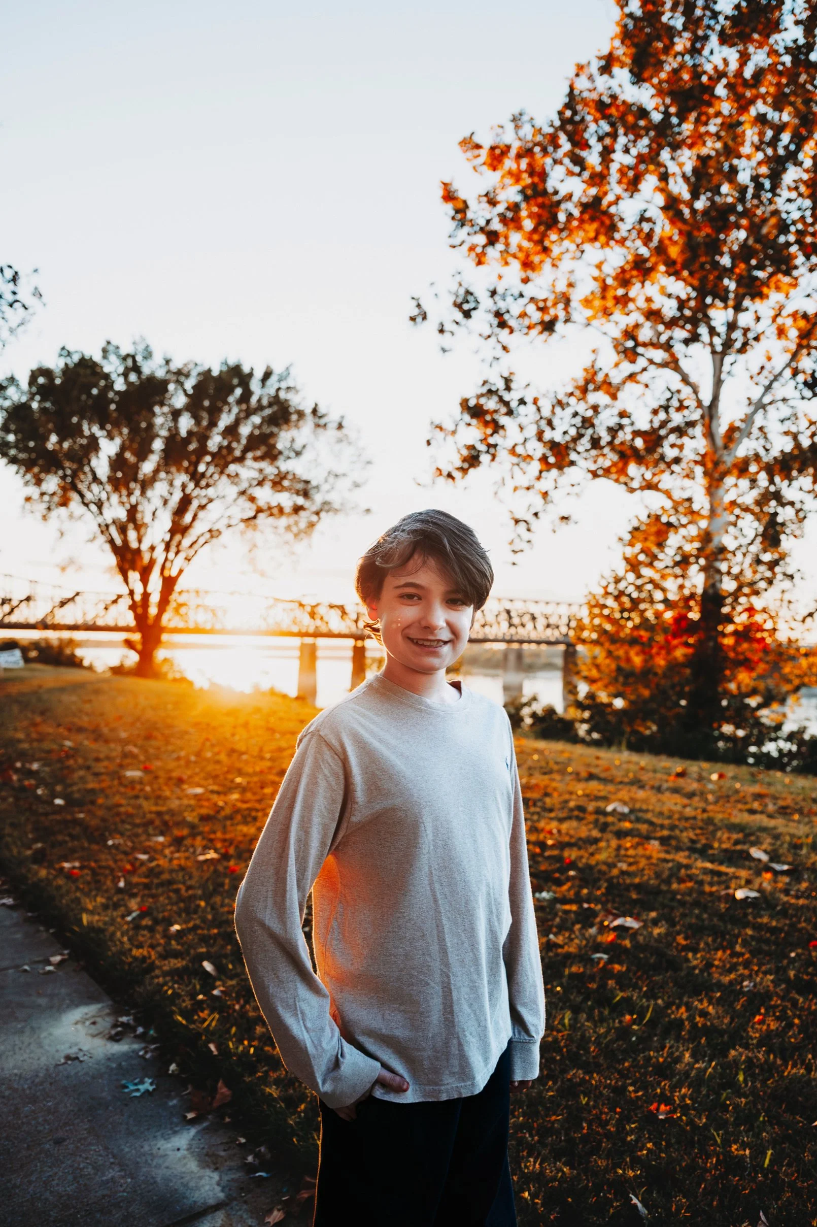 A young boy with dark hair smiling outdoors during sunset, standing on a grassy hill with trees and a bridge in the background.