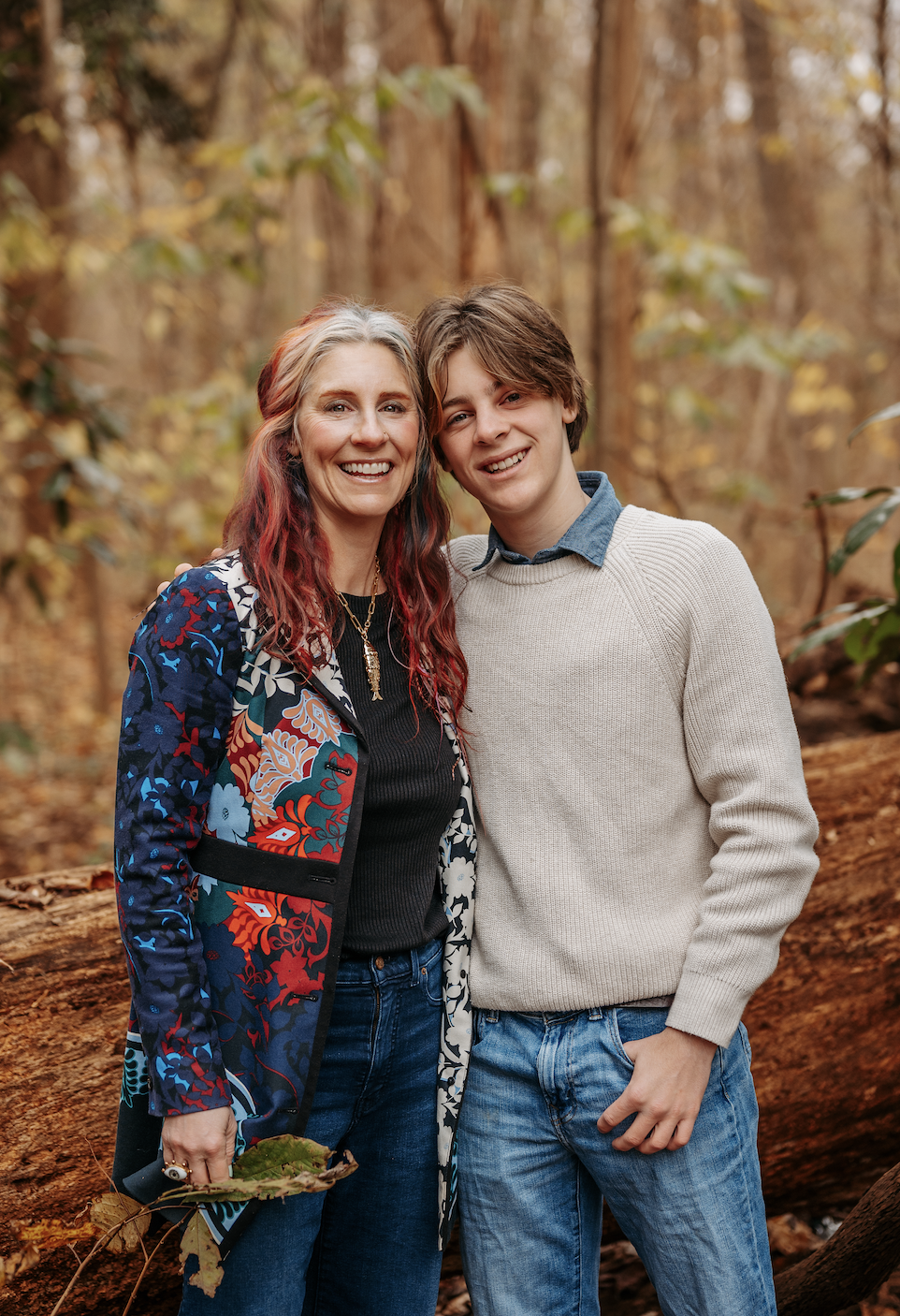 A smiling woman and young man standing close together in a forest with trees and fallen leaves, wearing casual clothes.