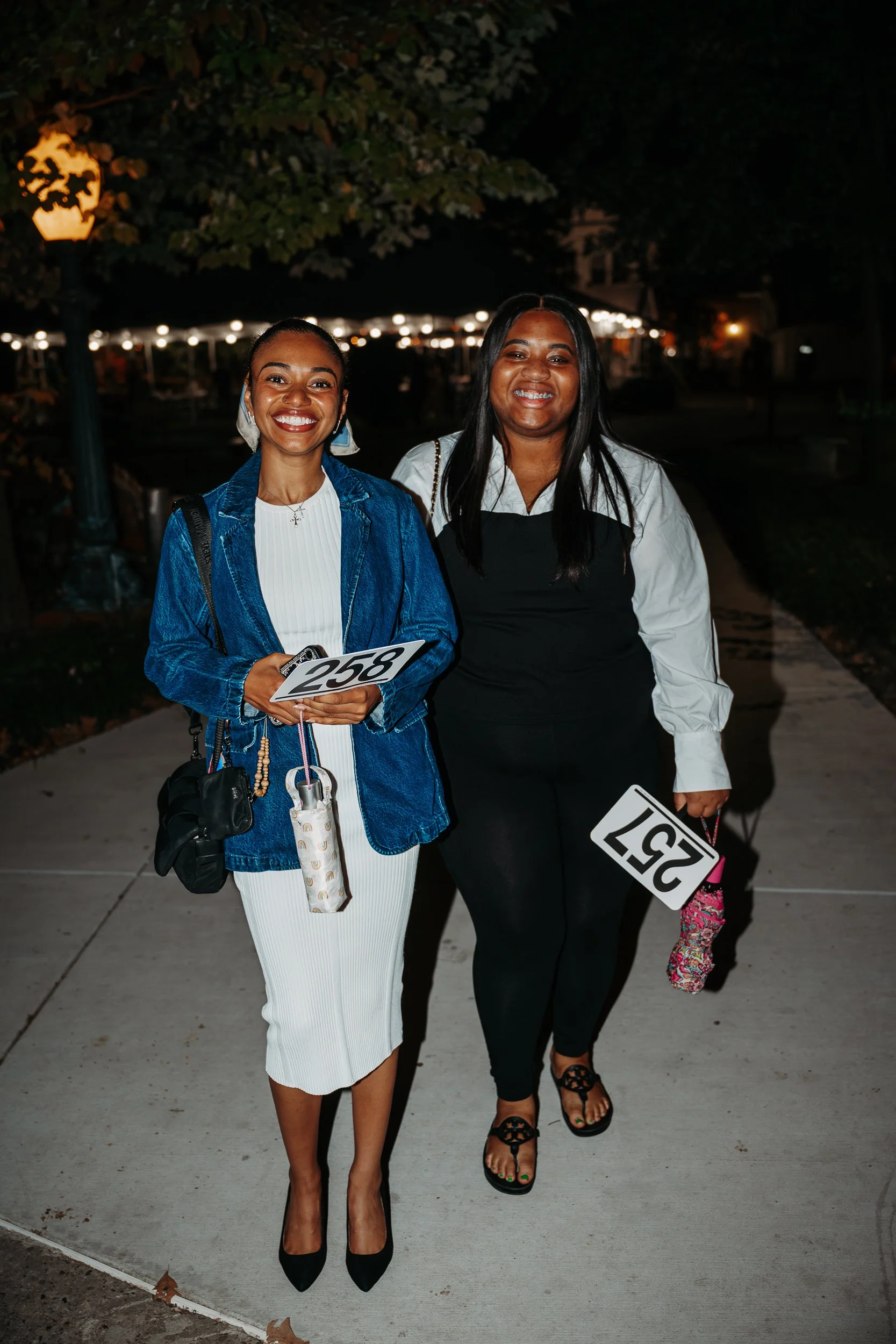 Two women standing on a sidewalk at night holding number signs, smiling at the camera.