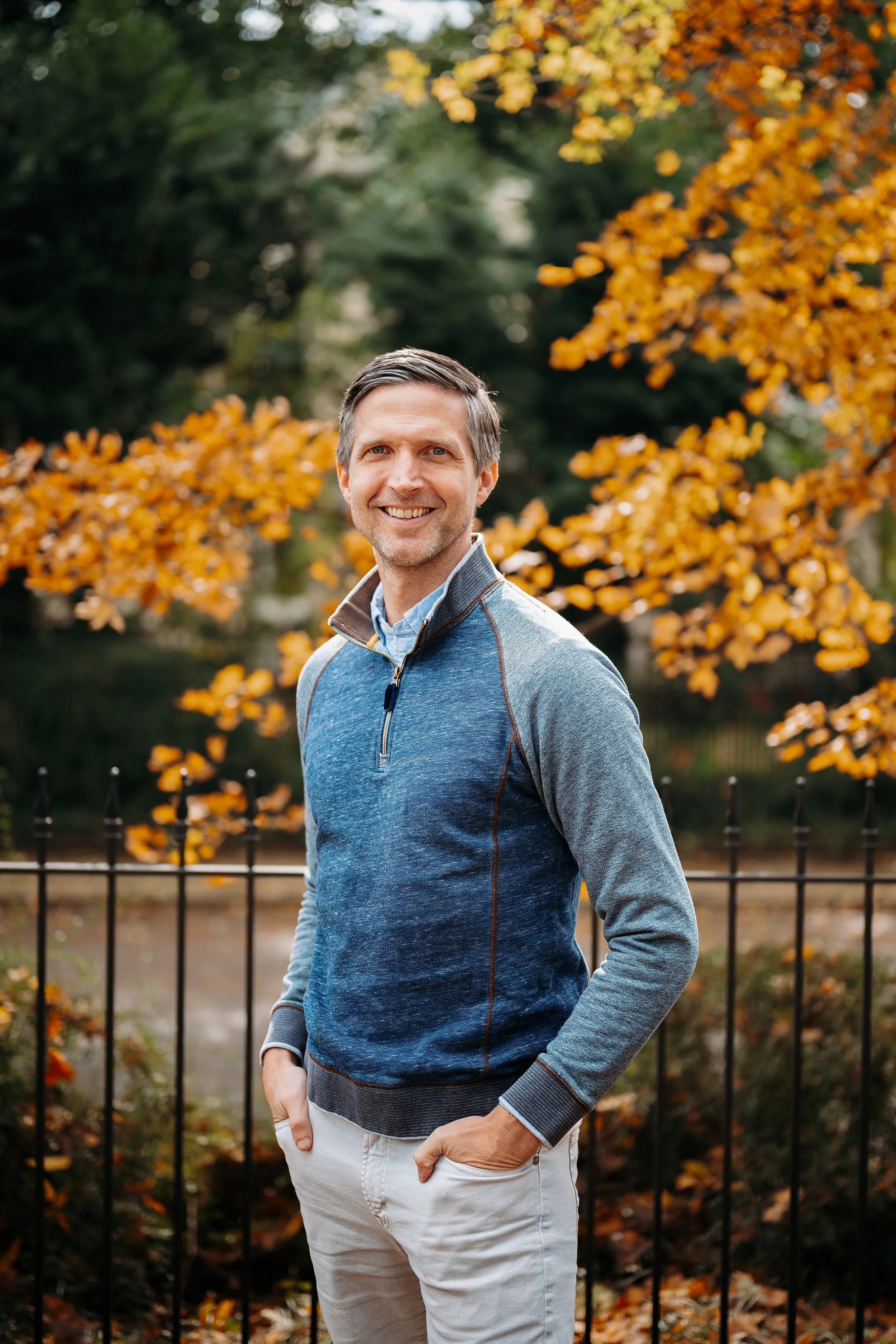 A man with gray hair and a beard smiling outdoors in front of a black iron fence and orange autumn leaves.