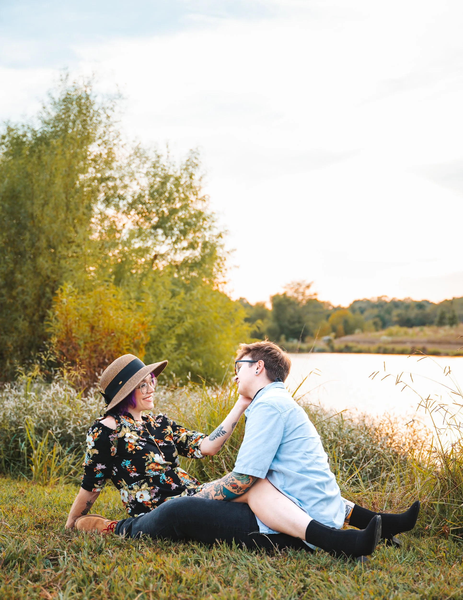 Two women sitting on the grass near a river, one with a hat and glasses, touching the other's face, during sunset.
