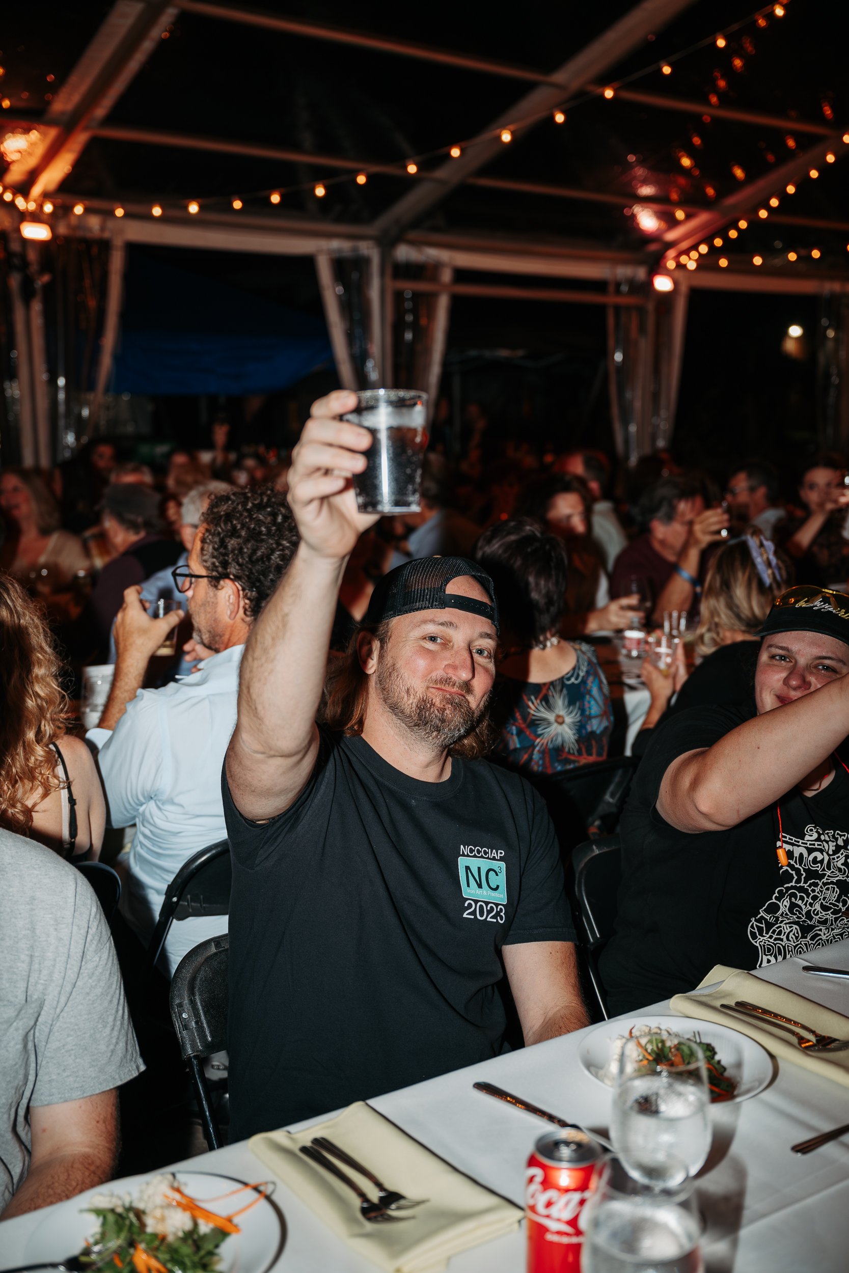 A man with long hair, a beard, and a black cap is smiling and raising a glass of water at a busy indoor event, surrounded by other people at a long table with plates, glasses, and silverware.