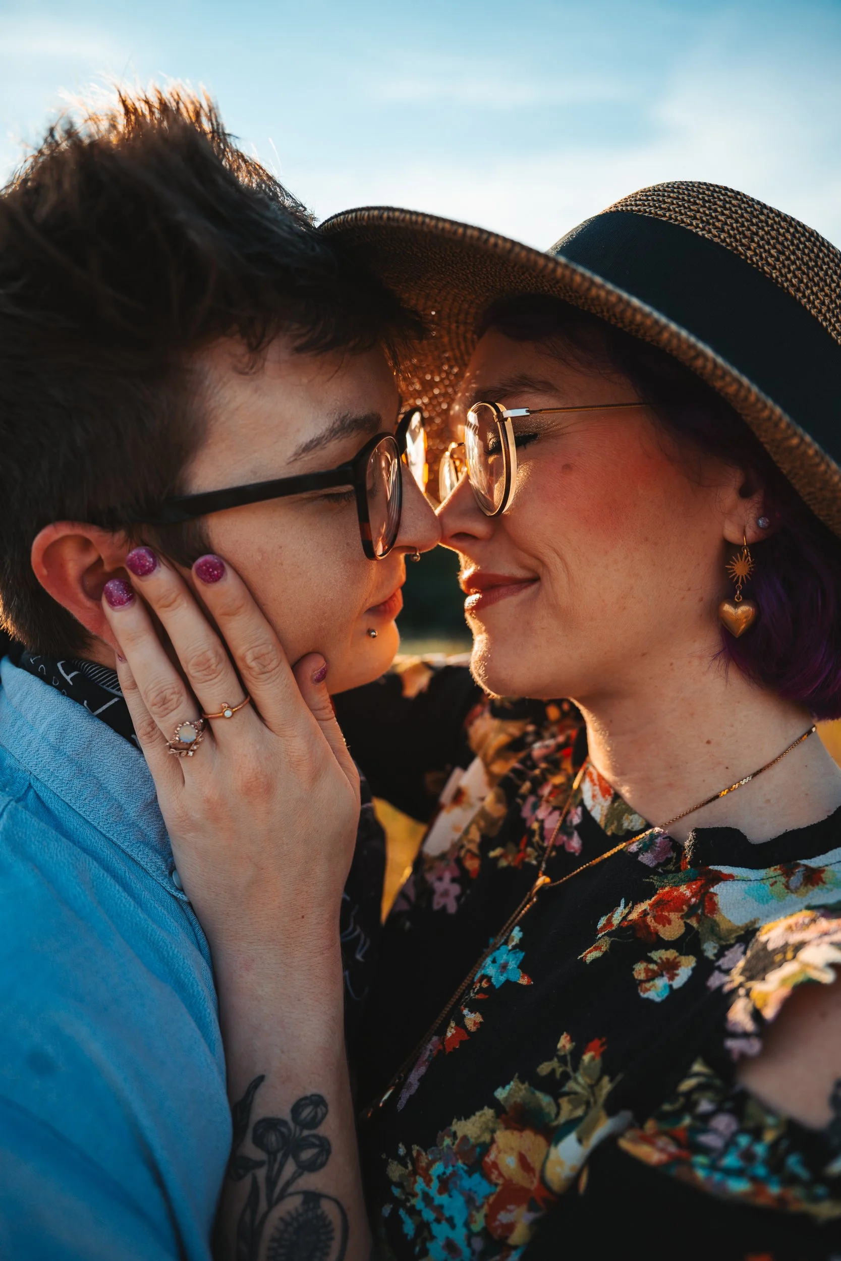 Two women are close together, touching foreheads and noses, smiling. One woman has short dark hair, glasses, a nose ring, and rings on her fingers. The other woman is wearing a large sunhat, glasses, earrings, and a floral top, holding the other woma