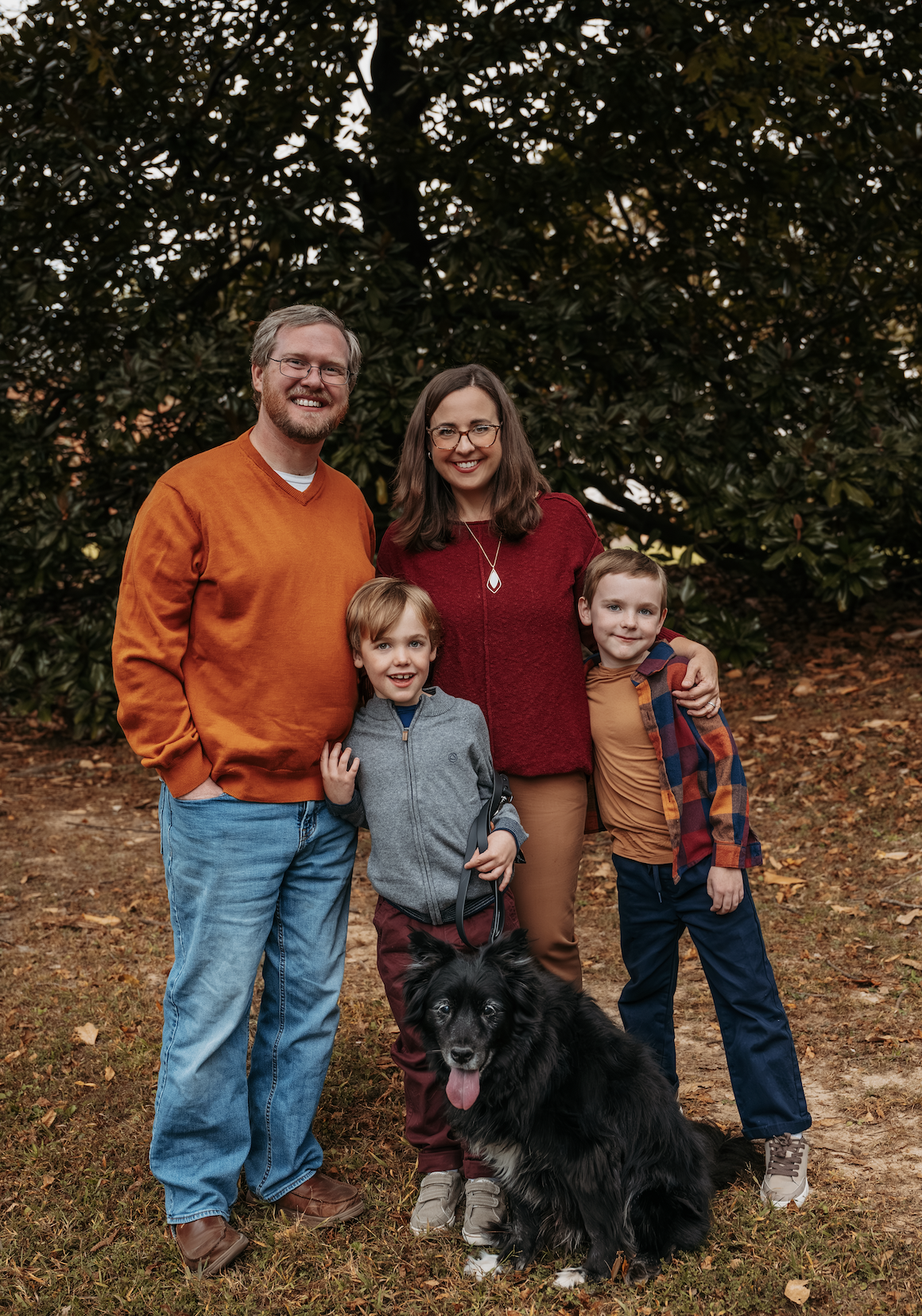 A family of four with two boys and a dog standing outdoors in front of trees, smiling at the camera during fall.