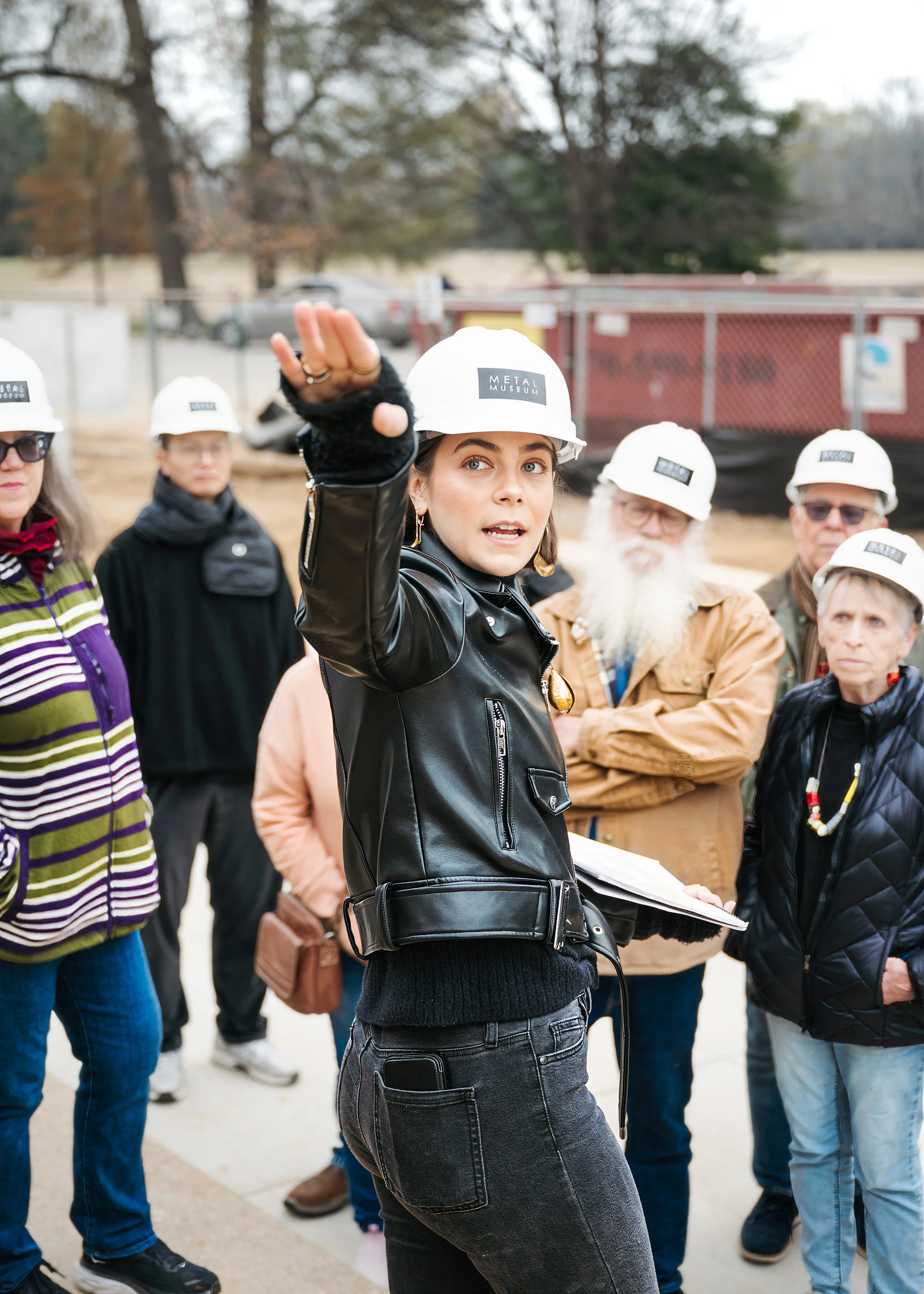 Young woman in a black leather jacket and white safety helmet speaking, with a group of diverse adults wearing similar safety helmets listening outdoors.