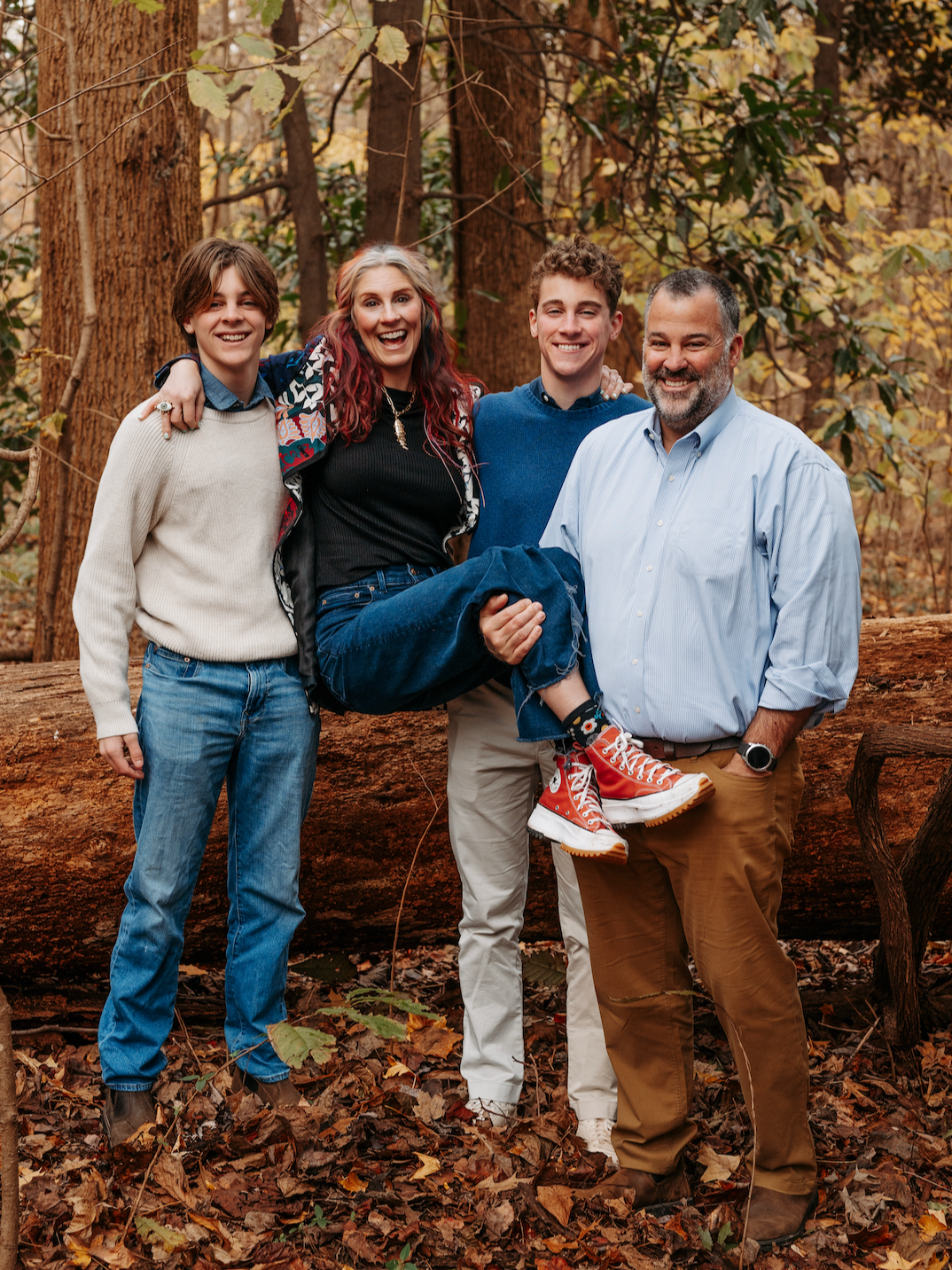 A family of four smiling and standing in a forest during fall, with three young adults and an older man, one woman being carried on the man's lap.