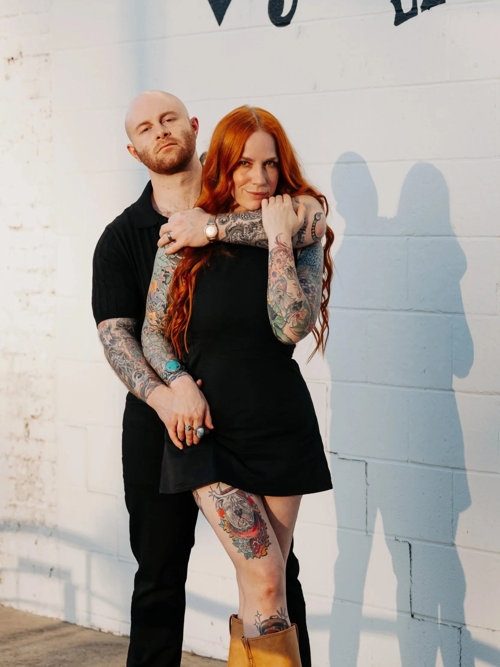 A man and woman with tattoos standing close together against a white brick wall, casting shadows on the wall.