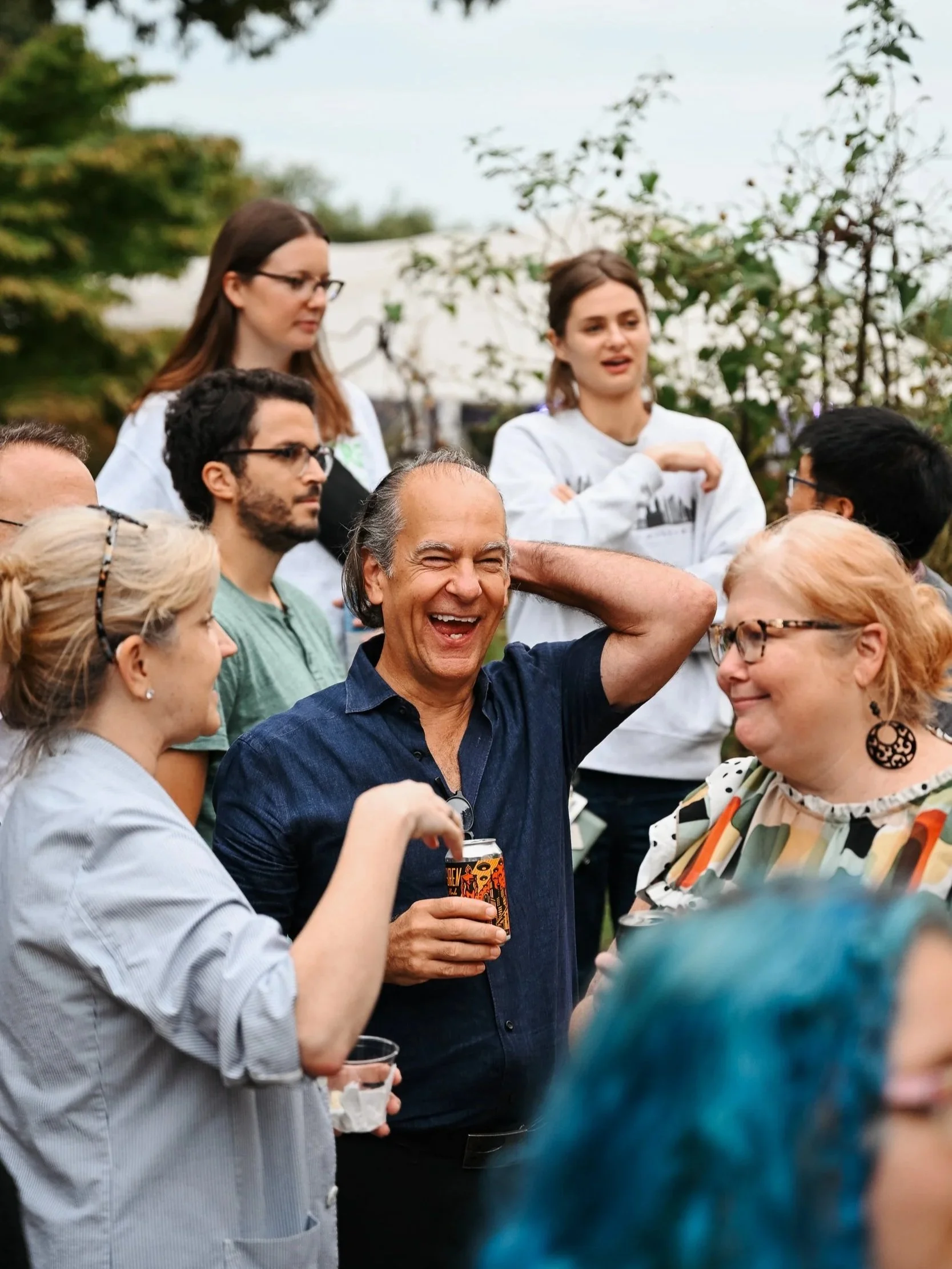 Group of diverse people socializing outdoors, smiling, and laughing at a gathering, with trees and a tent in the background.