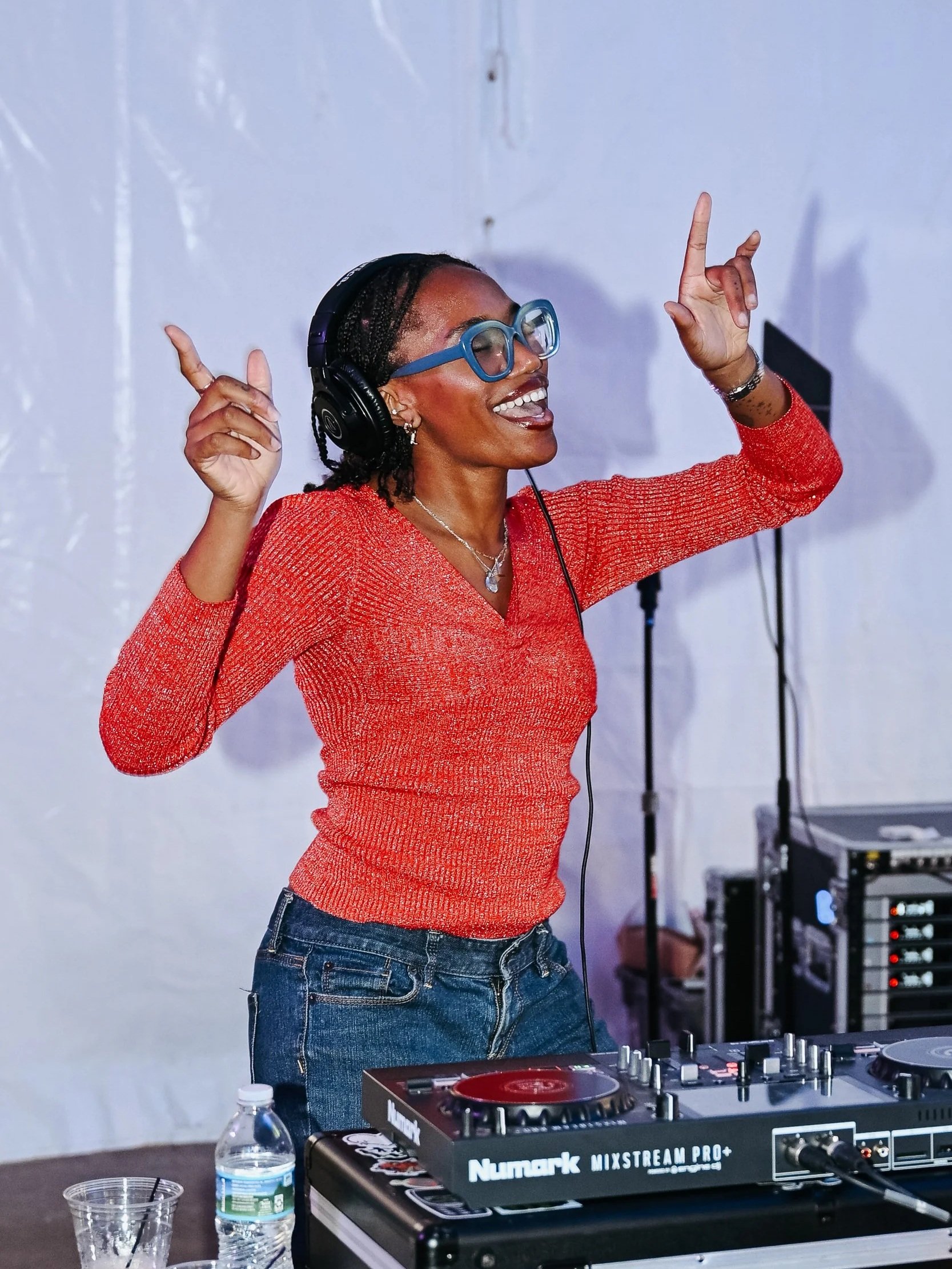 A woman with glasses and headphones dancing and smiling behind DJ equipment.