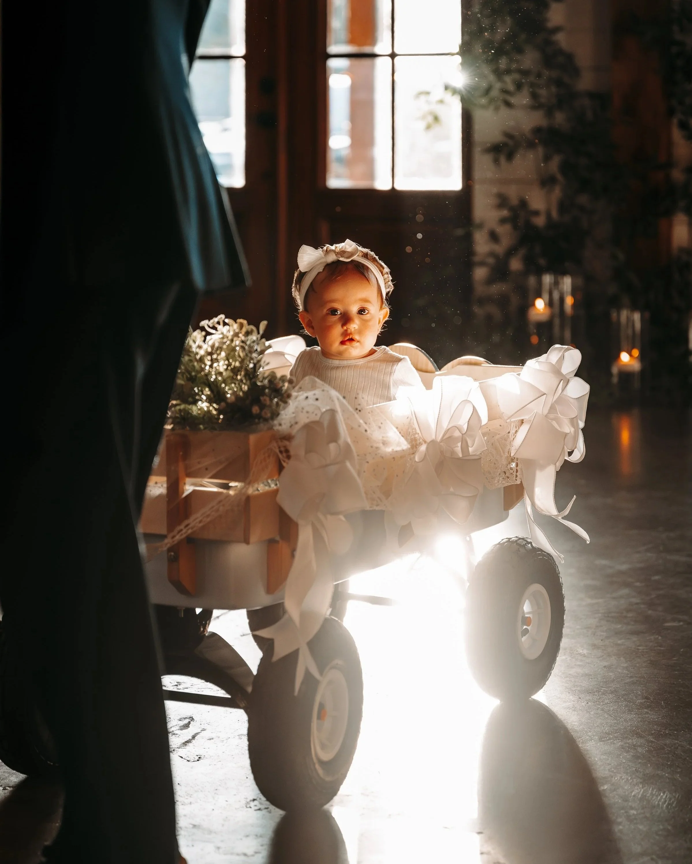 A young child sitting in a decorated wagon with white bows and flowers inside, illuminated by sunlight through windows in the background, with candles on the side.