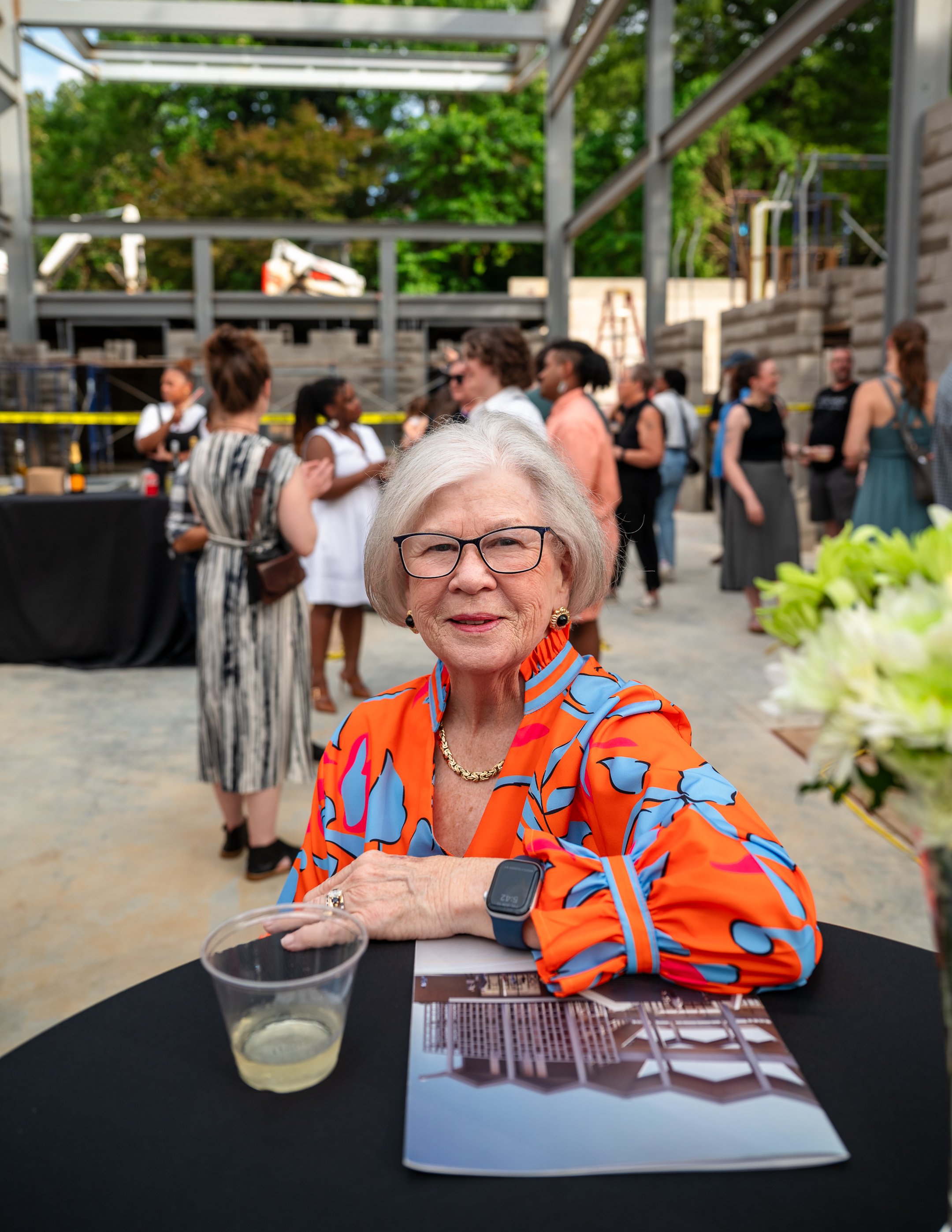 An elderly woman with glasses, wearing an orange and blue patterned jacket, sitting at a black table with a drink, a magazine, and a smartwatch, at a social gathering with multiple people in the background.