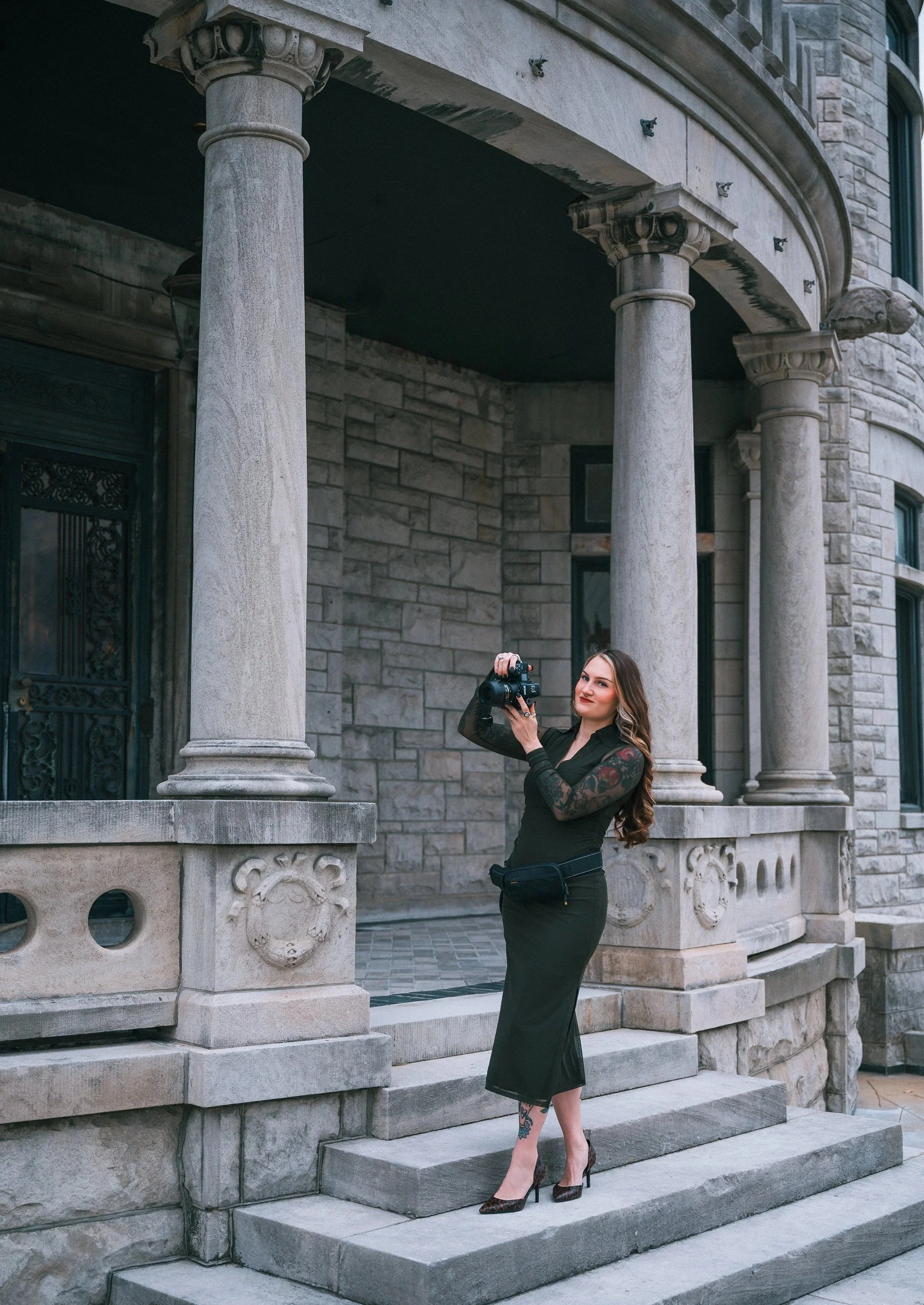 A woman in a black dress and high heels standing on the steps outside a large stone building, holding a camera and smiling.