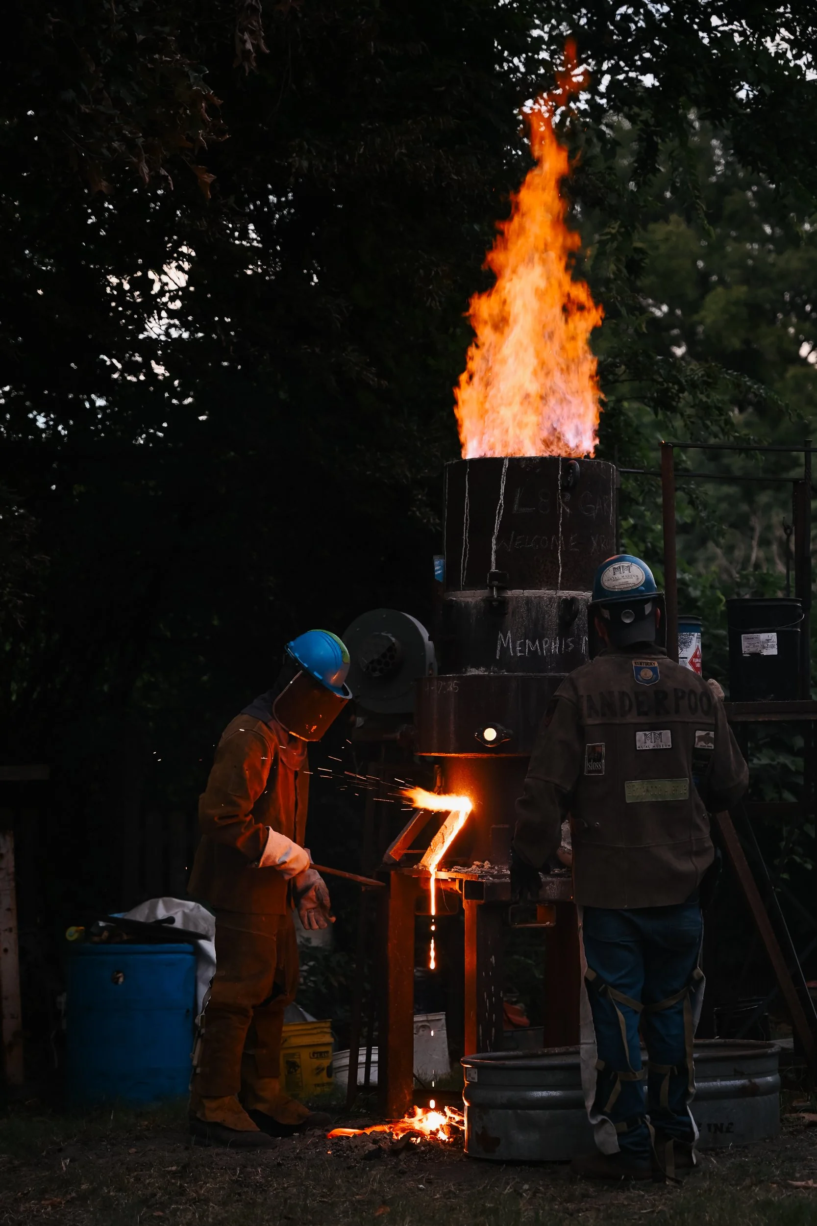 Two metalworkers are pouring molten metal into a mold outdoors, with a large flame erupting from the furnace behind them. They are wearing protective gear, including helmets and gloves.