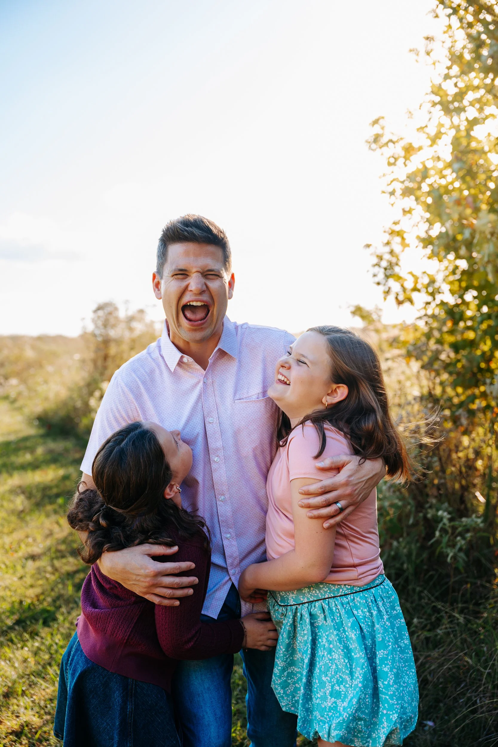 A man and two young girls outdoors, smiling and hugging each other, with trees and a clear sky in the background.