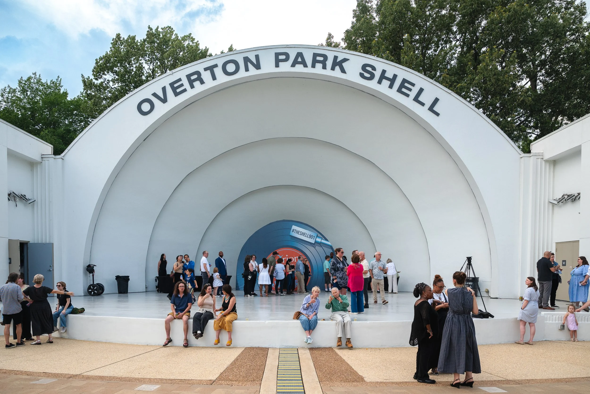 People gather on an outdoor stage at Overton Park Shell during daytime, with some sitting on the edge of the stage. The stage has a white semi-circular shell structure with 