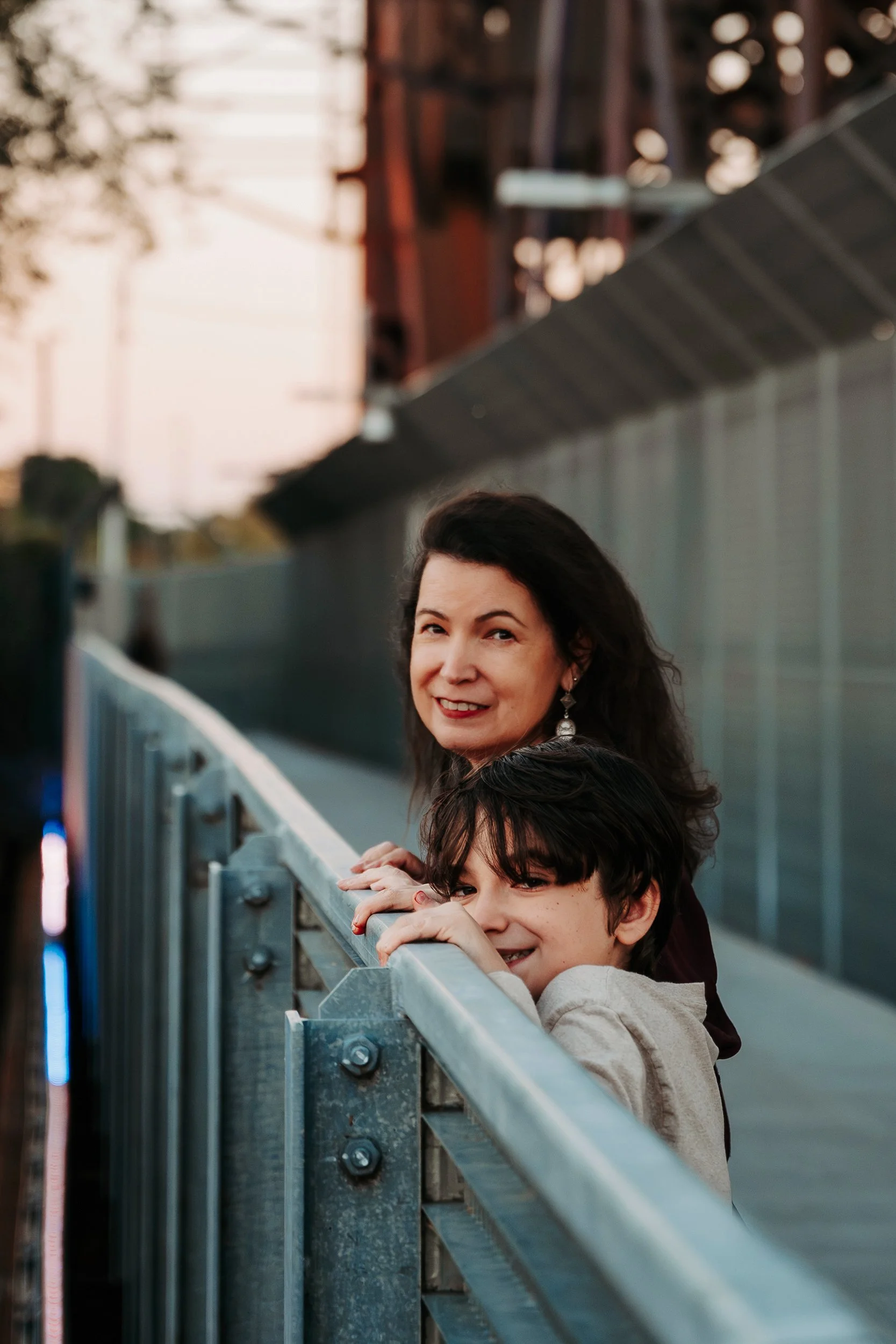 A woman and a young boy leaning on a metal railing on a bridge at sunset, smiling and looking at the camera.
