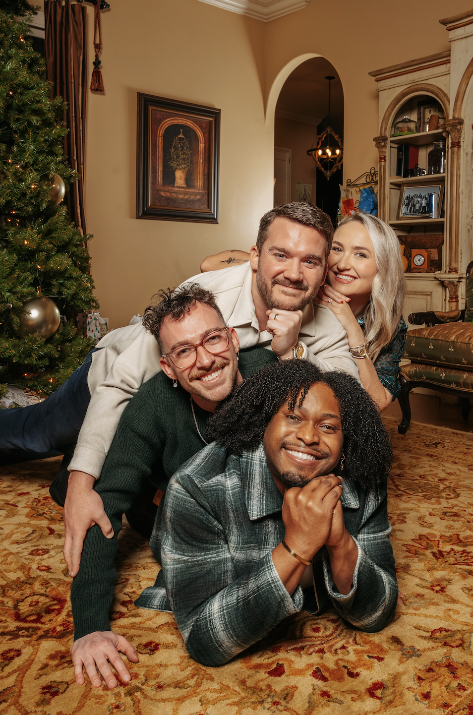 Celebrating Christmas with four friends, smiling and lying on a decorated living room rug near a Christmas tree with gold ornaments. Around 2023.