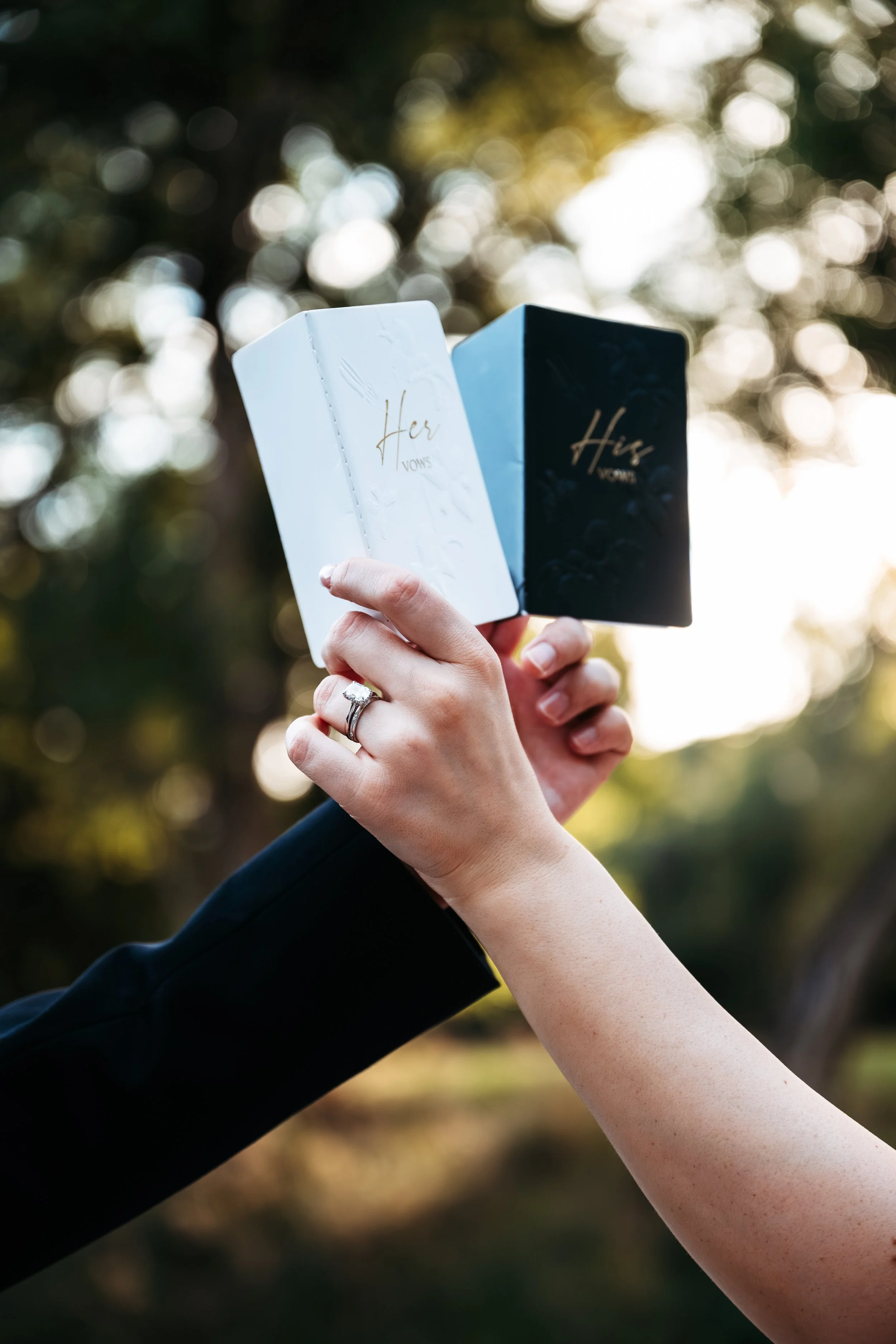 A person holding two small prayer books, one white and one black, outdoors with blurred trees and sunlight in the background.