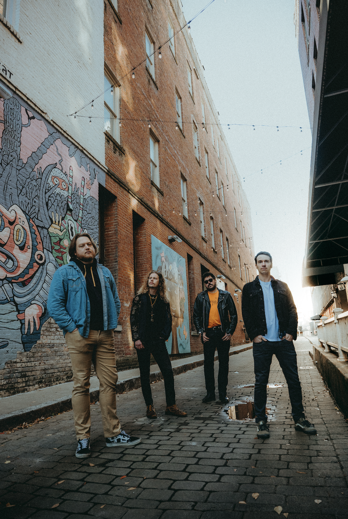 Four people standing on a cobblestone street with brick buildings and colorful murals, daytime with string lights above.