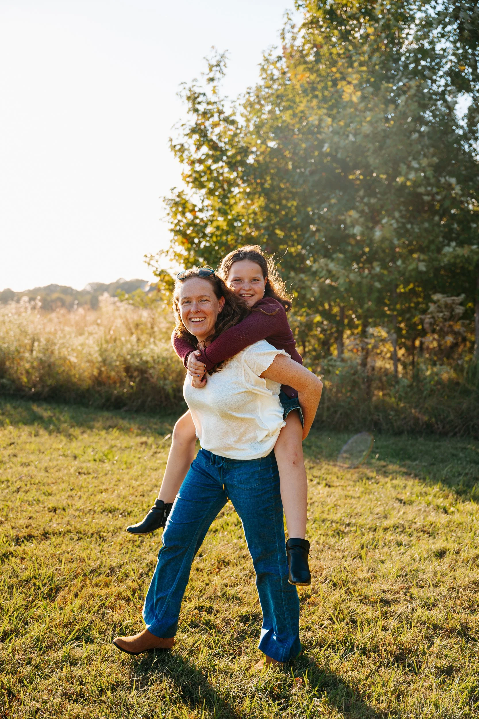 A woman giving a piggyback ride to a girl in a field during golden hour, with trees in the background.