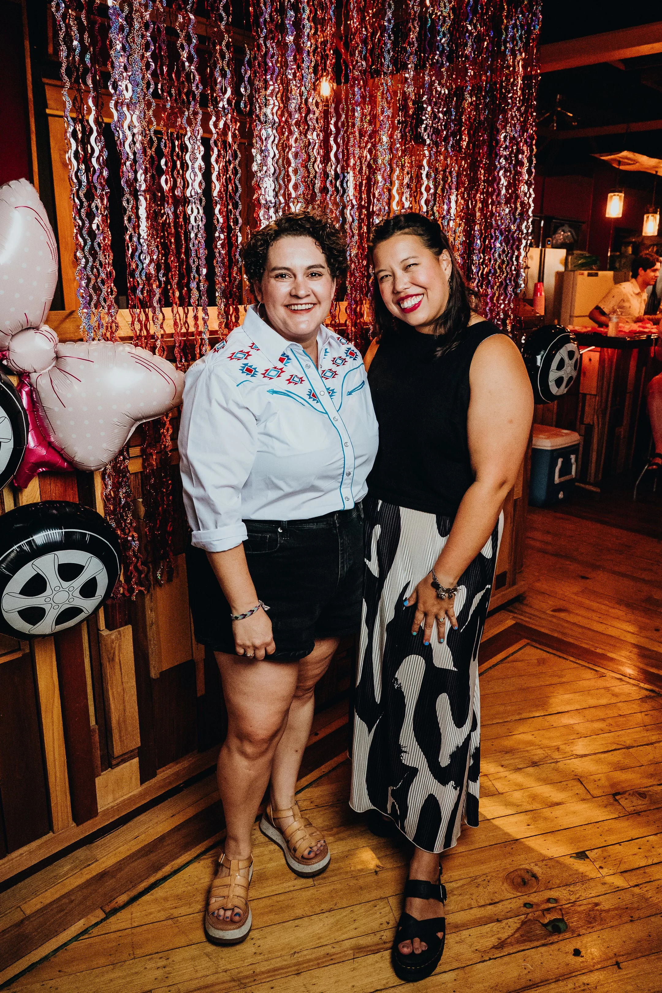 Two women smiling at a party with pink and purple hanging decorations behind them.