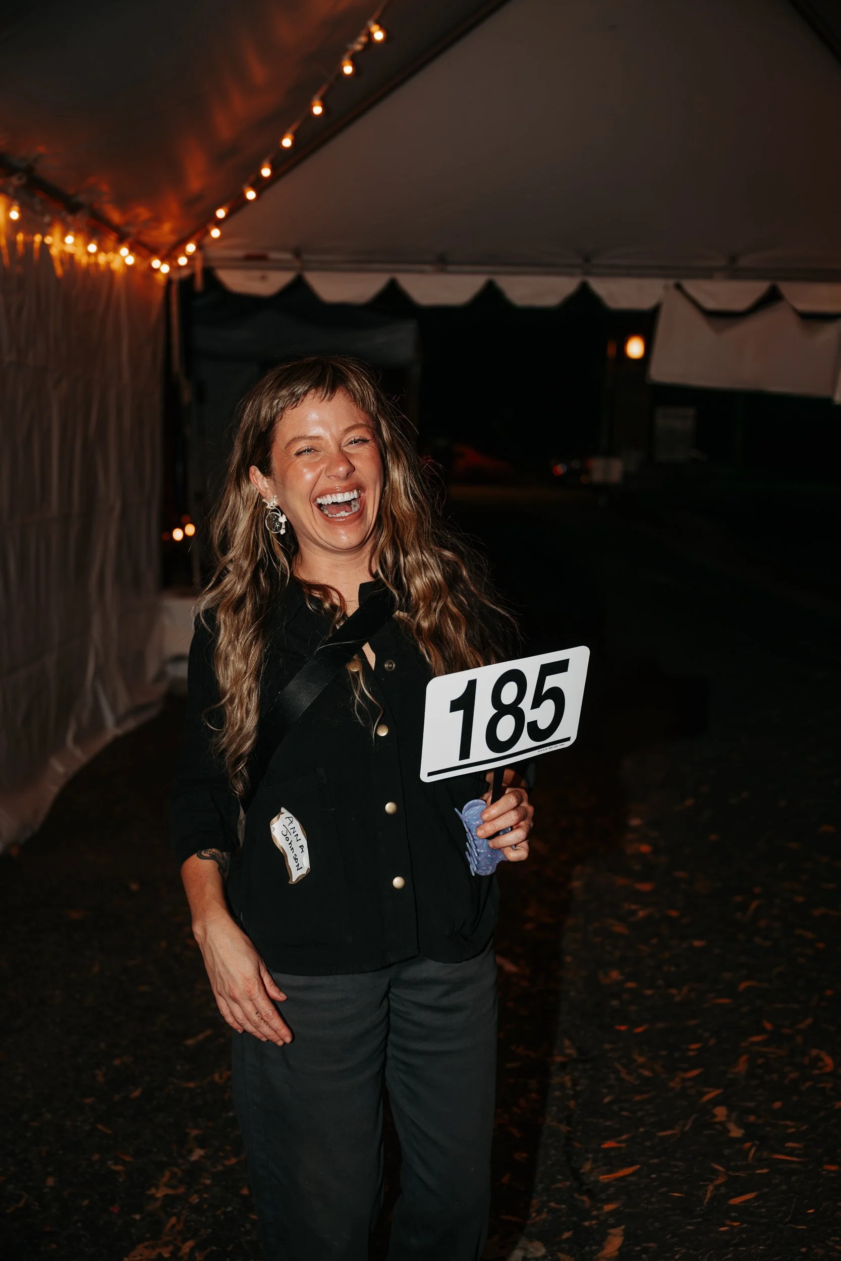 A woman with long curly hair wearing earrings and a black shirt, smiling and laughing, holding a sign with the number 185, standing under a tent with string lights at night.