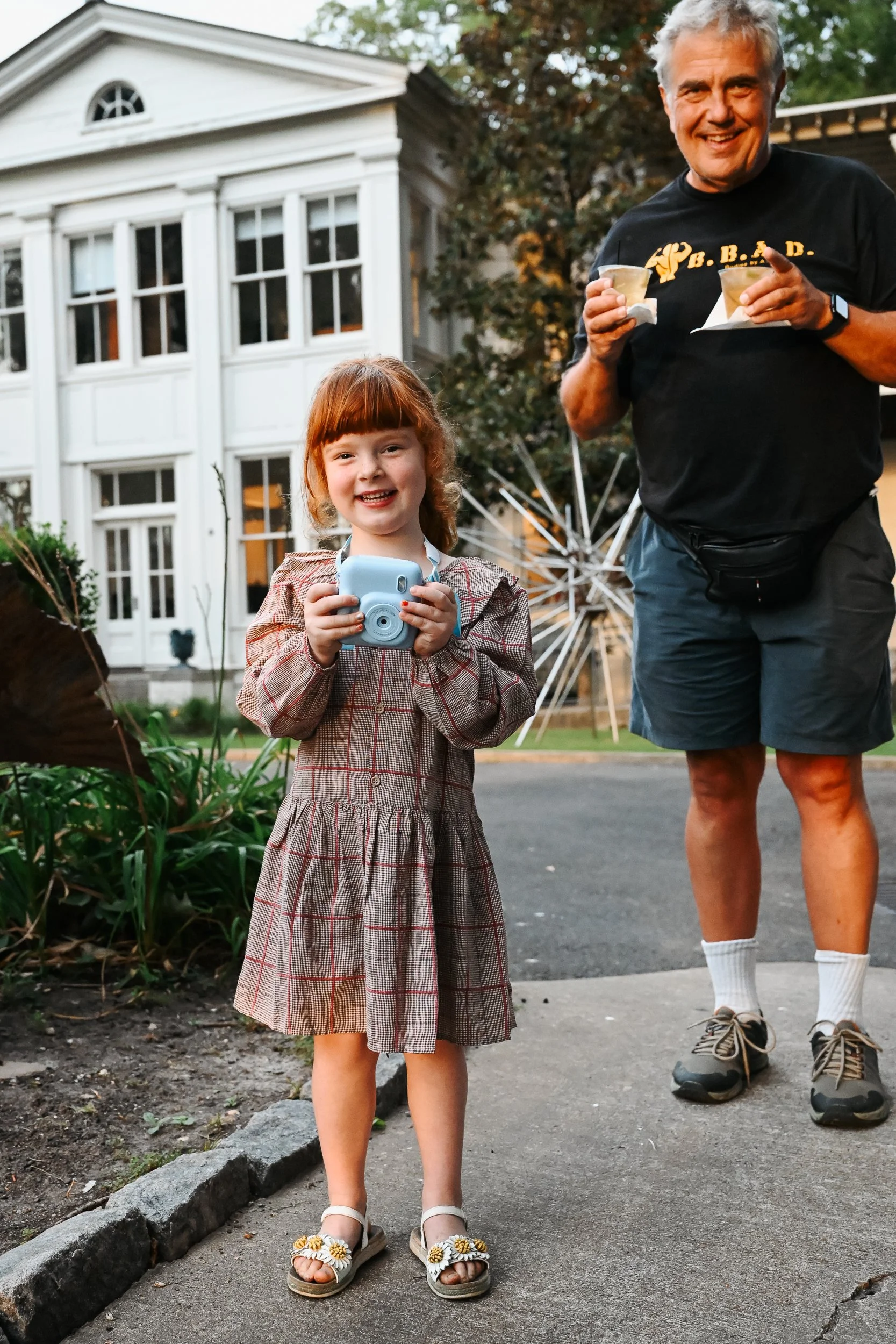 A young girl with red hair in a plaid dress holding a camera and smiling, standing outdoors on a sidewalk with a man in shorts behind her, both near a white house with large windows and a garden with plants and a large white outdoor sculpture in the 