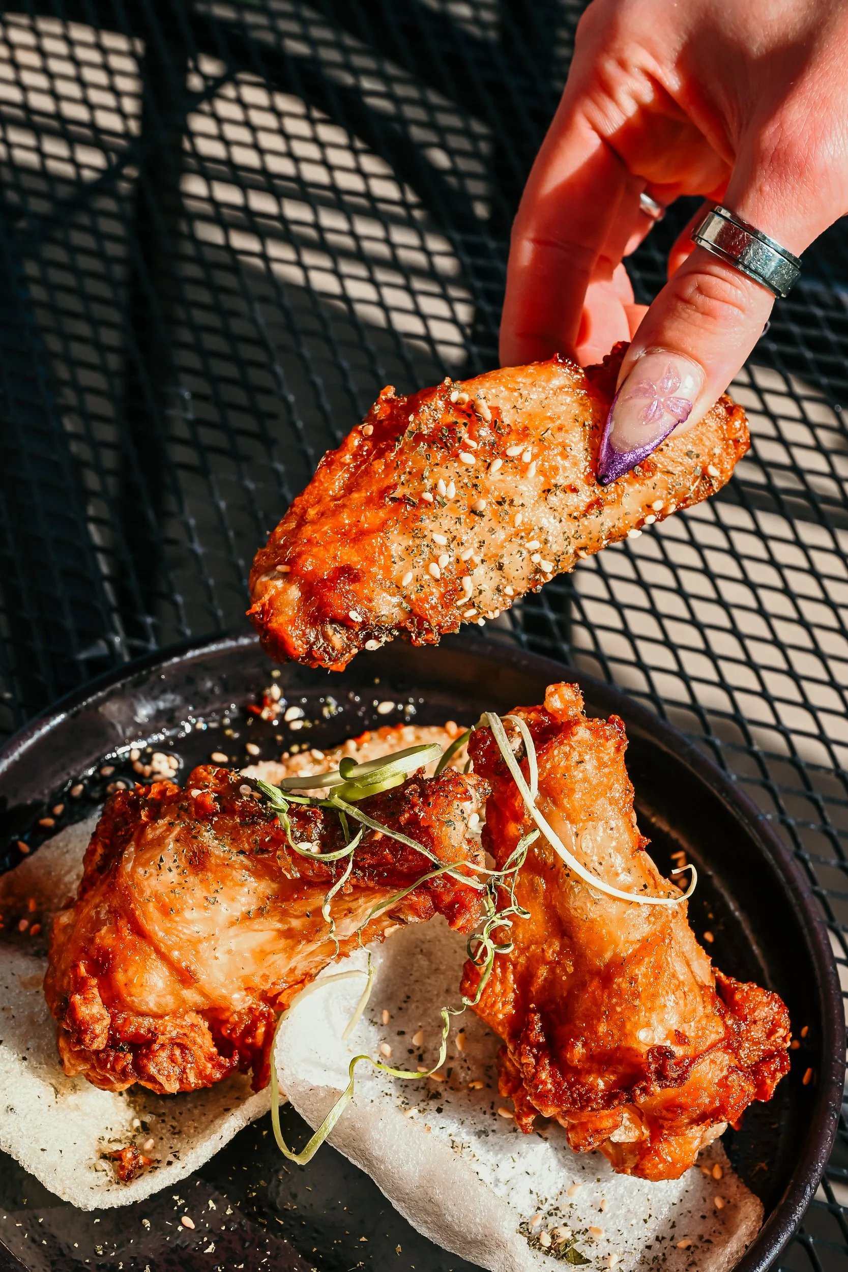Person grabbing a piece of fried chicken seasoned with herbs and sesame seeds from a plate containing additional pieces of fried chicken garnished with microgreens and served on a black metal outdoor table.