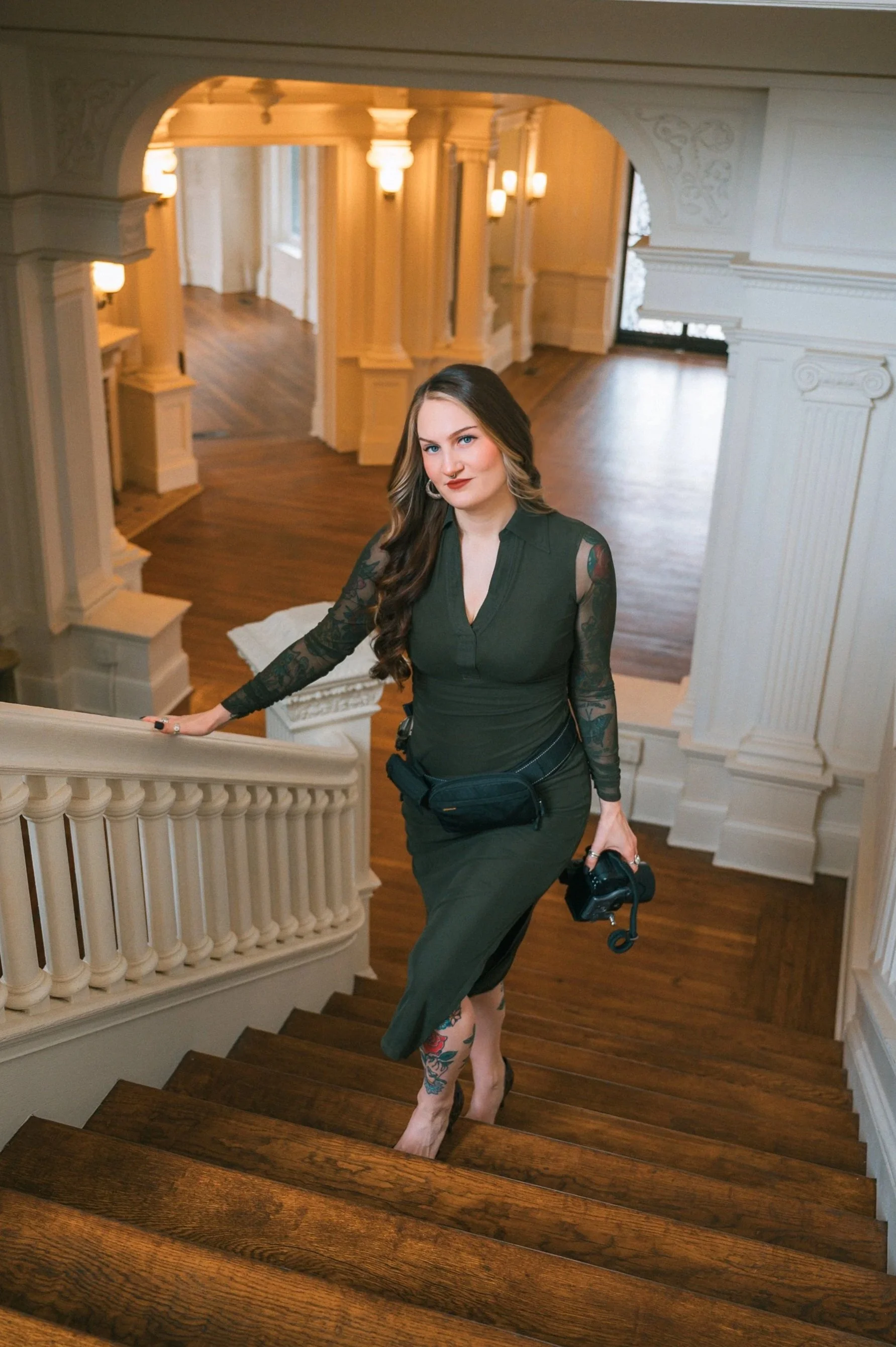 A woman with long, wavy hair, wearing a dark green dress with lace sleeves, standing on a wooden staircase inside a grand, well-lit building with ornate white walls and columns, holding a camera and a camera strap.