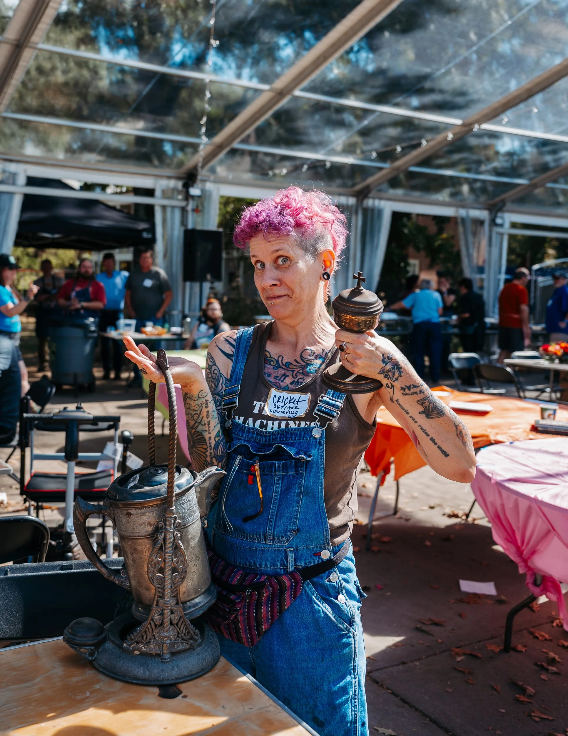 A woman with pink curly hair, tattoos, and wearing overalls holds a decorative chalice in one hand and a vintage-style teapot in the other, standing at an outdoor event with tables and people in the background.