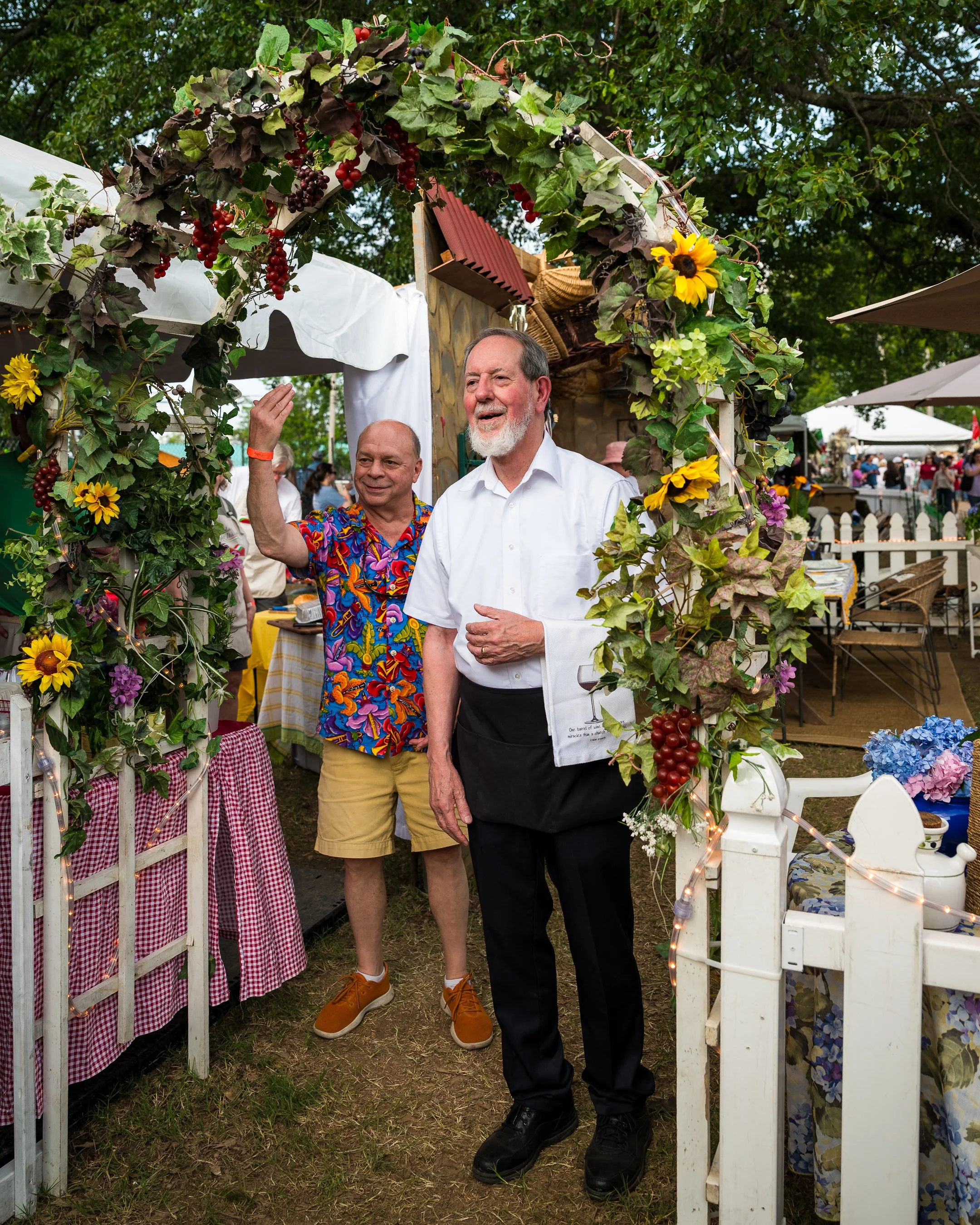 Two men standing behind a decorated arch at an outdoor event, surrounded by tables, chairs, and other people.