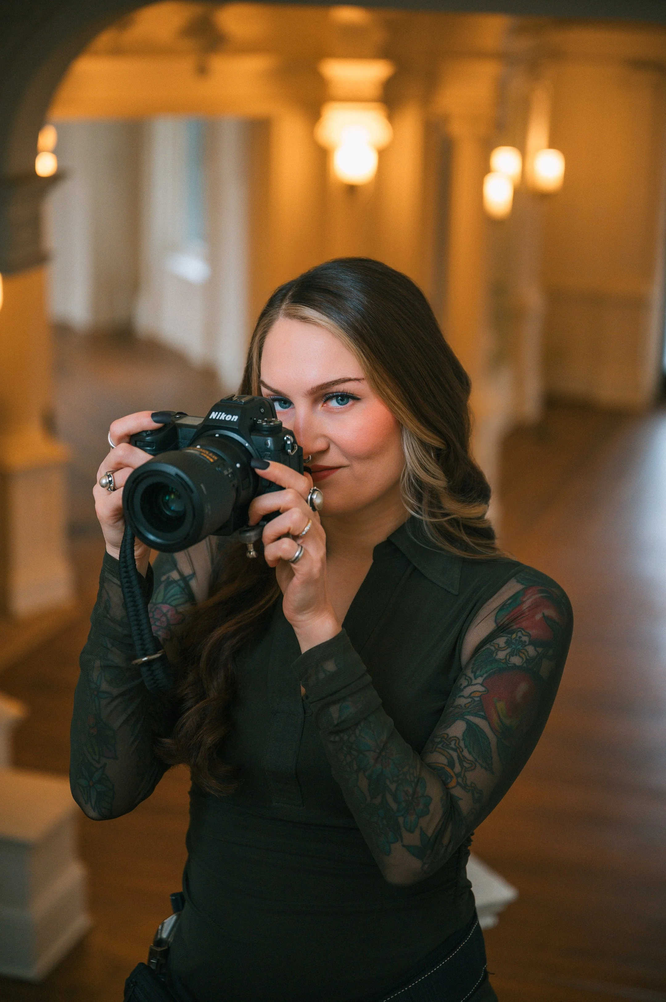 A woman with long brown hair and tattoos on her arms, dressed in black, aims a Nikon camera directly at the viewer with a focused expression. She is standing indoors with warm lighting and blurred background.