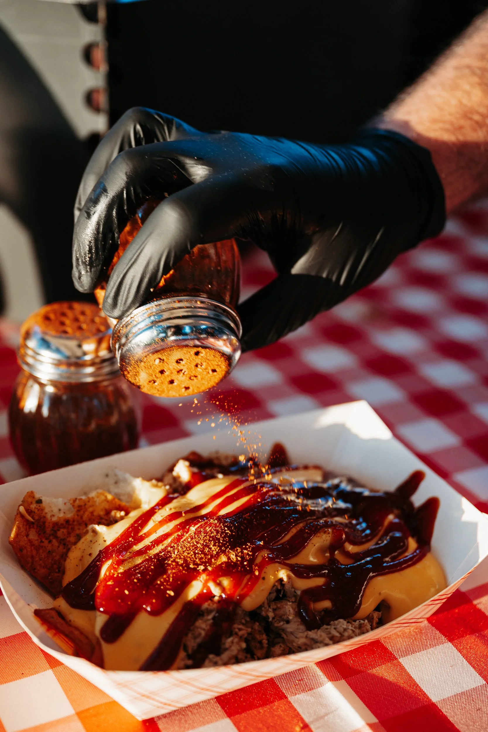 A person wearing a black glove sprinkling orange seasoning onto a serving of loaded fries topped with ketchup, mayonnaise, and barbecue sauce on a paper tray placed on a red and white checkered tablecloth.