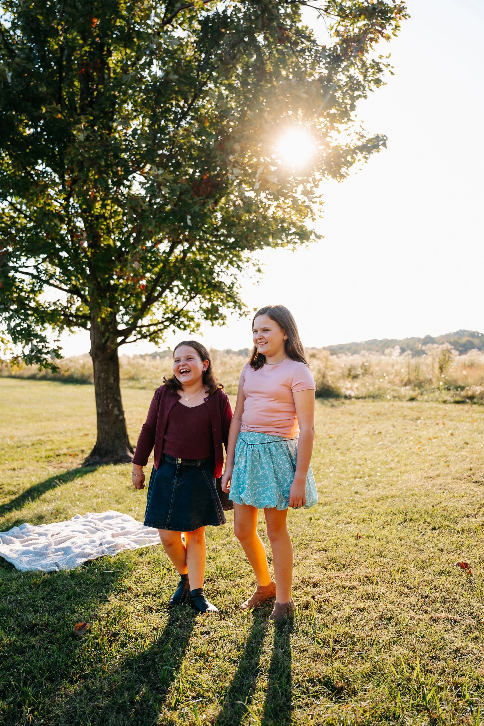 Two young girls standing outdoors in a grassy park area, smiling and laughing, with a large tree and the sun shining through the leaves in the background, during a sunny day.