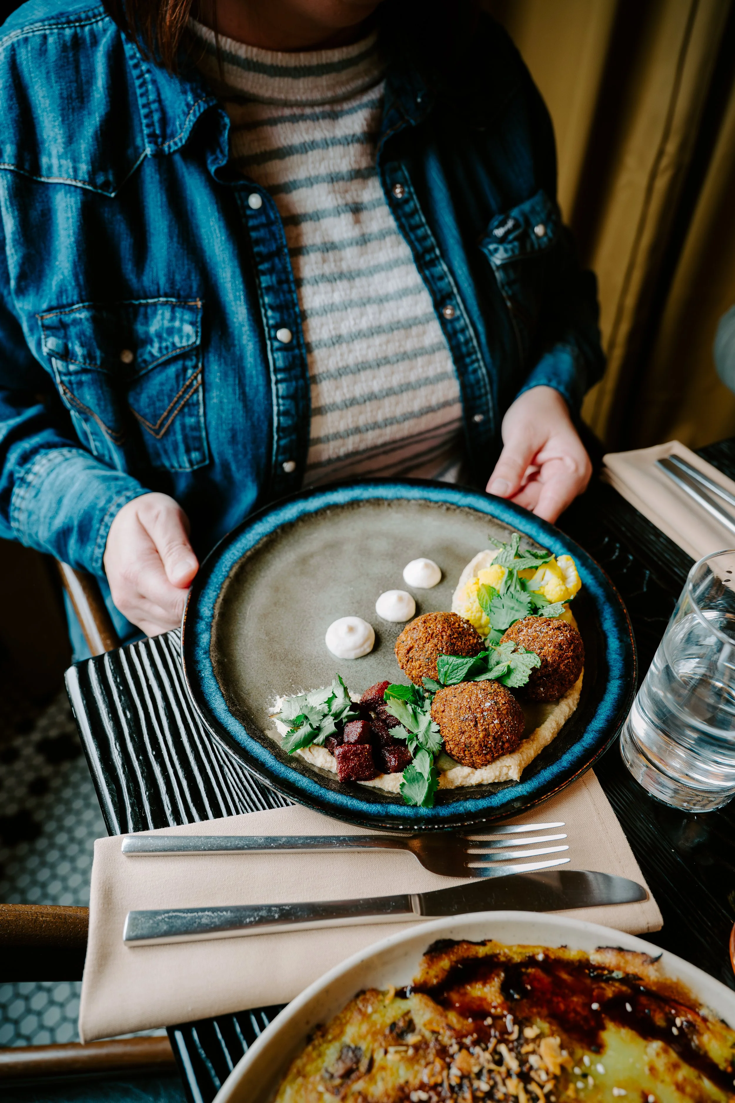 Close-up of a person wearing a denim jacket and striped shirt holding a plate with falafel balls, pickled vegetables, and herbs, on a dining table.